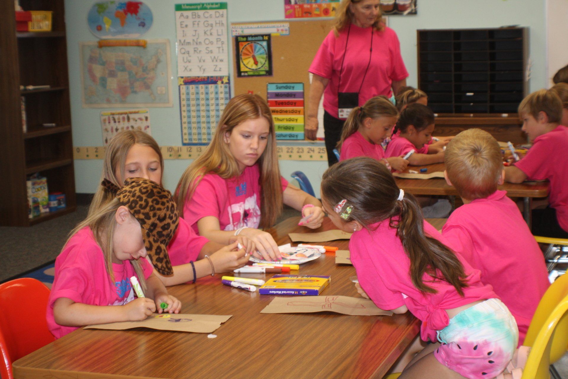 Children in pink shirts working at a classroom table while a teacher stands nearby