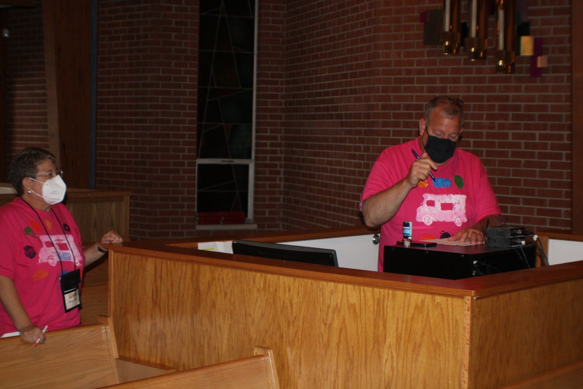 Two masked speakers in pink shirts stand at a church podium, one speaking into a microphone.