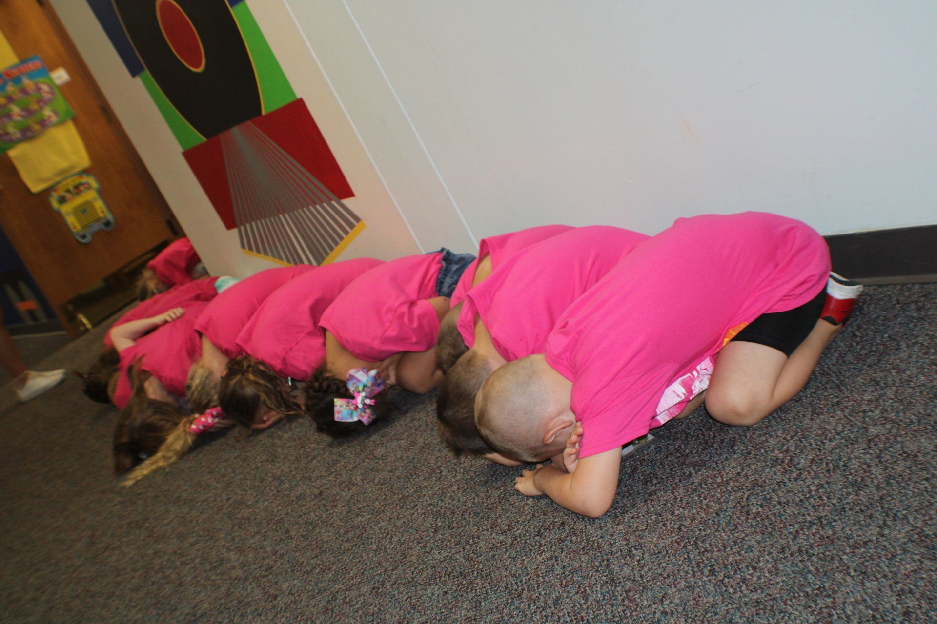 Several children in bright pink outfits crouch on a carpeted floor indoors, heads down in a group.