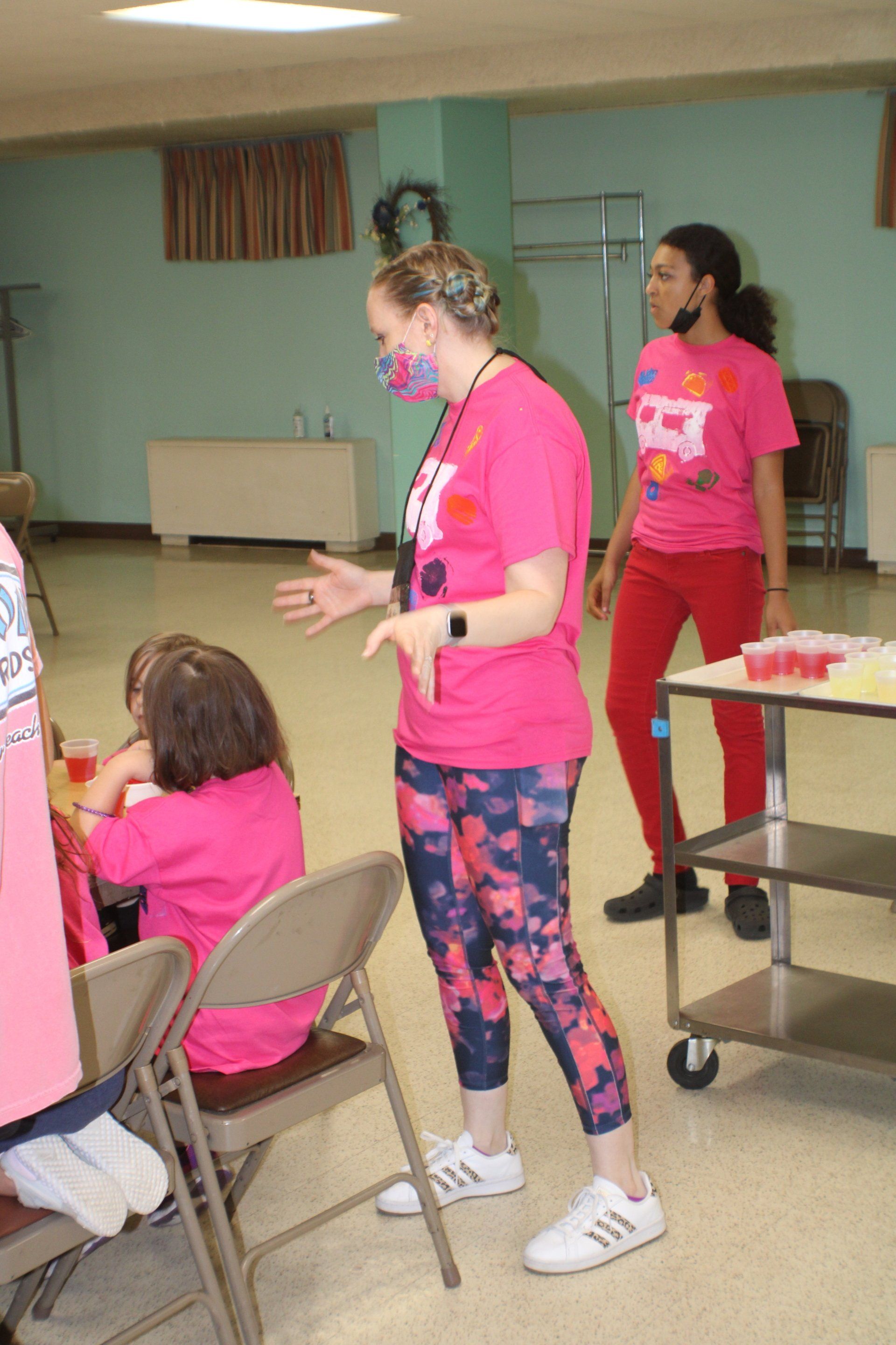 People in pink shirts gathered in a room, one standing and speaking near a seated group.