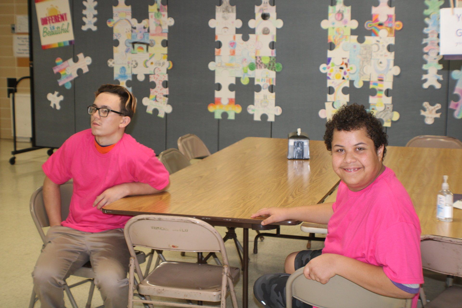 Two people in pink shirts seated at a table in a classroom with puzzle-piece wall decorations