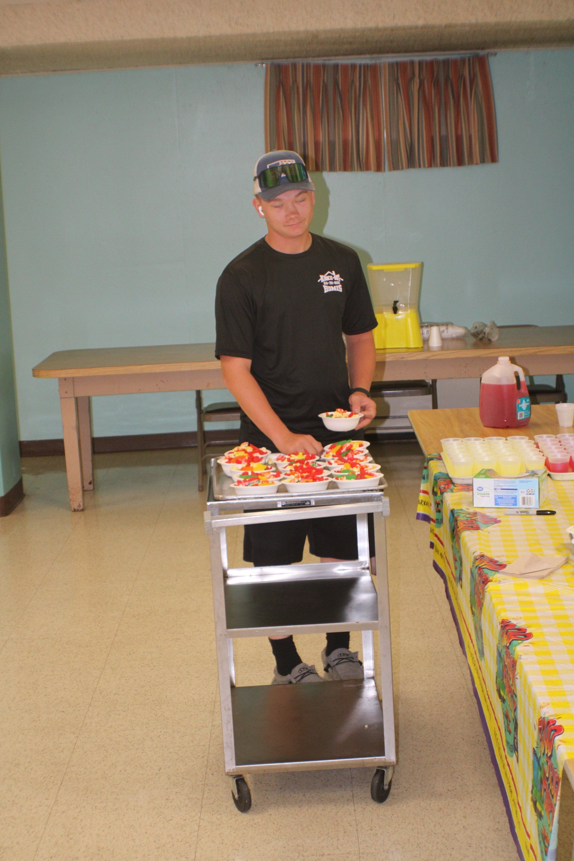 Man standing behind a serving cart with food in a room, beside a table covered with yellow cloth.