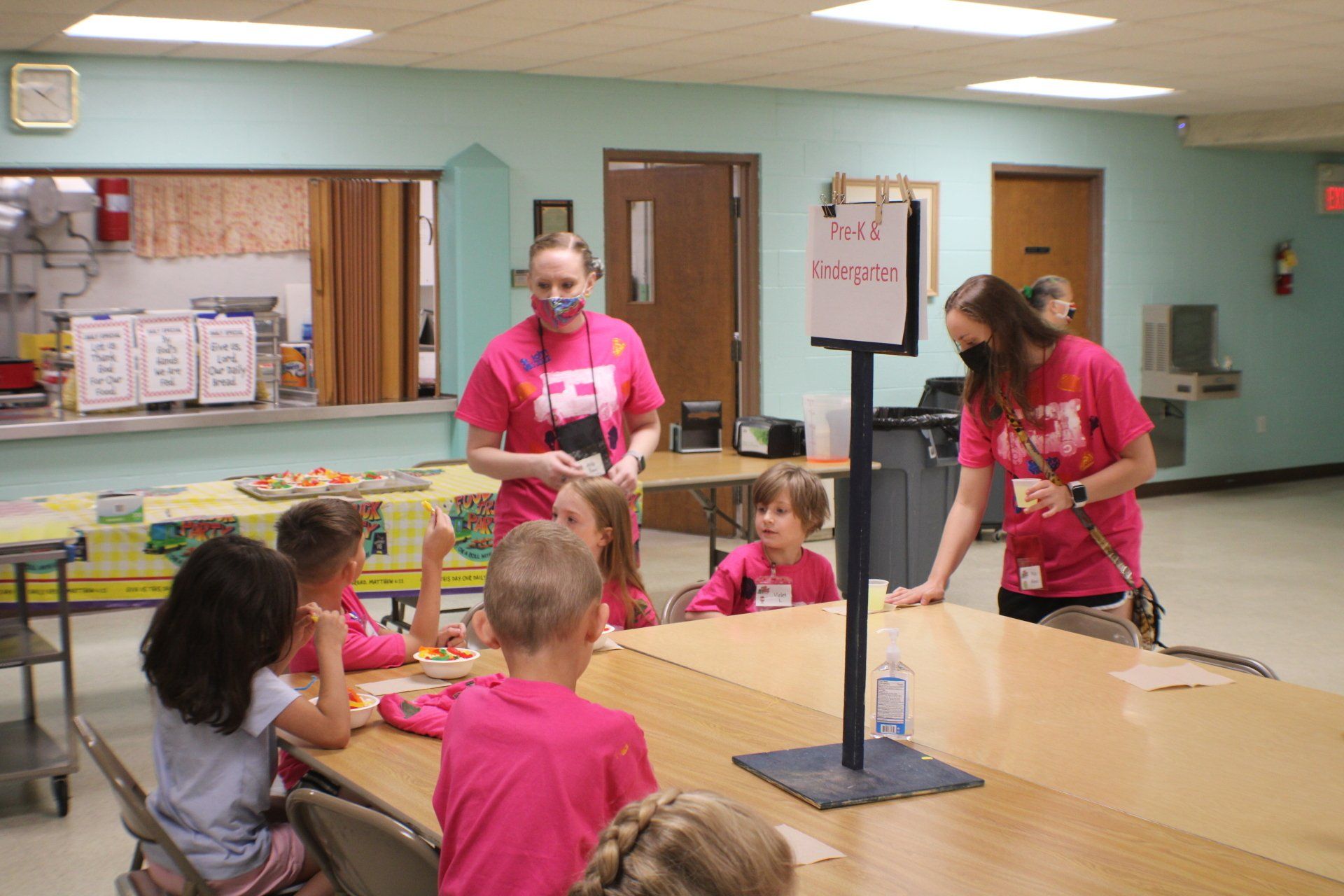 Children and adults in pink shirts gather around a table in a classroom or daycare room.