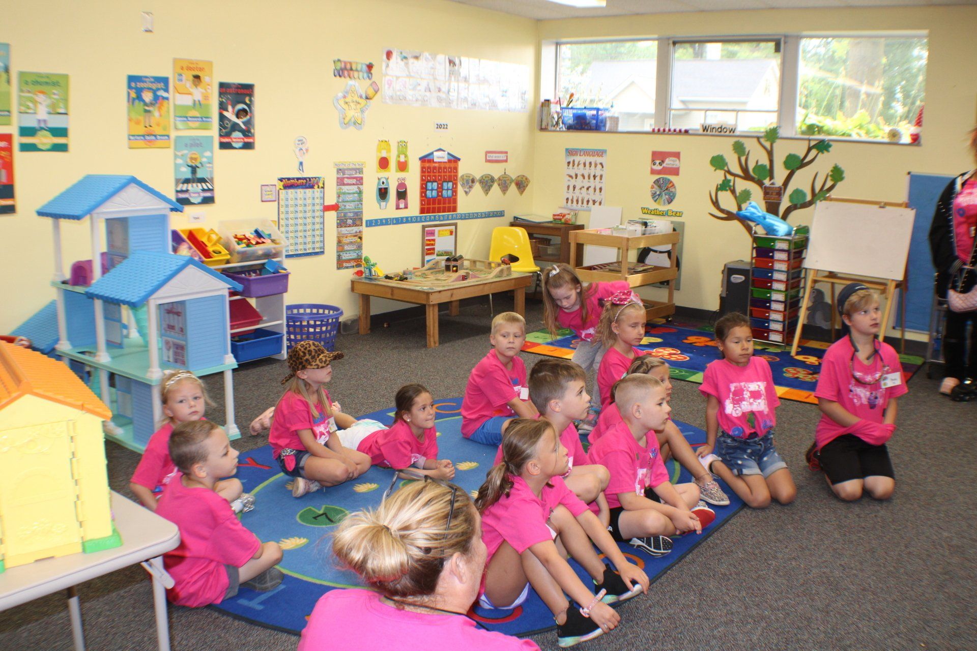 Children in pink outfits seated in a bright classroom, listening to a teacher near a toy display.