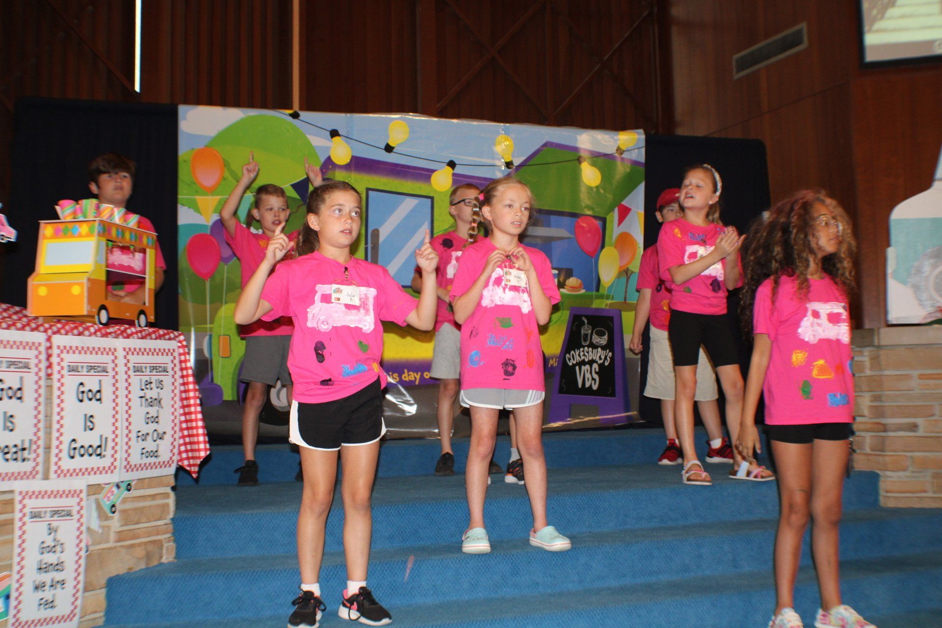 Children in pink shirts performing on a stage with colorful cartoon backdrop