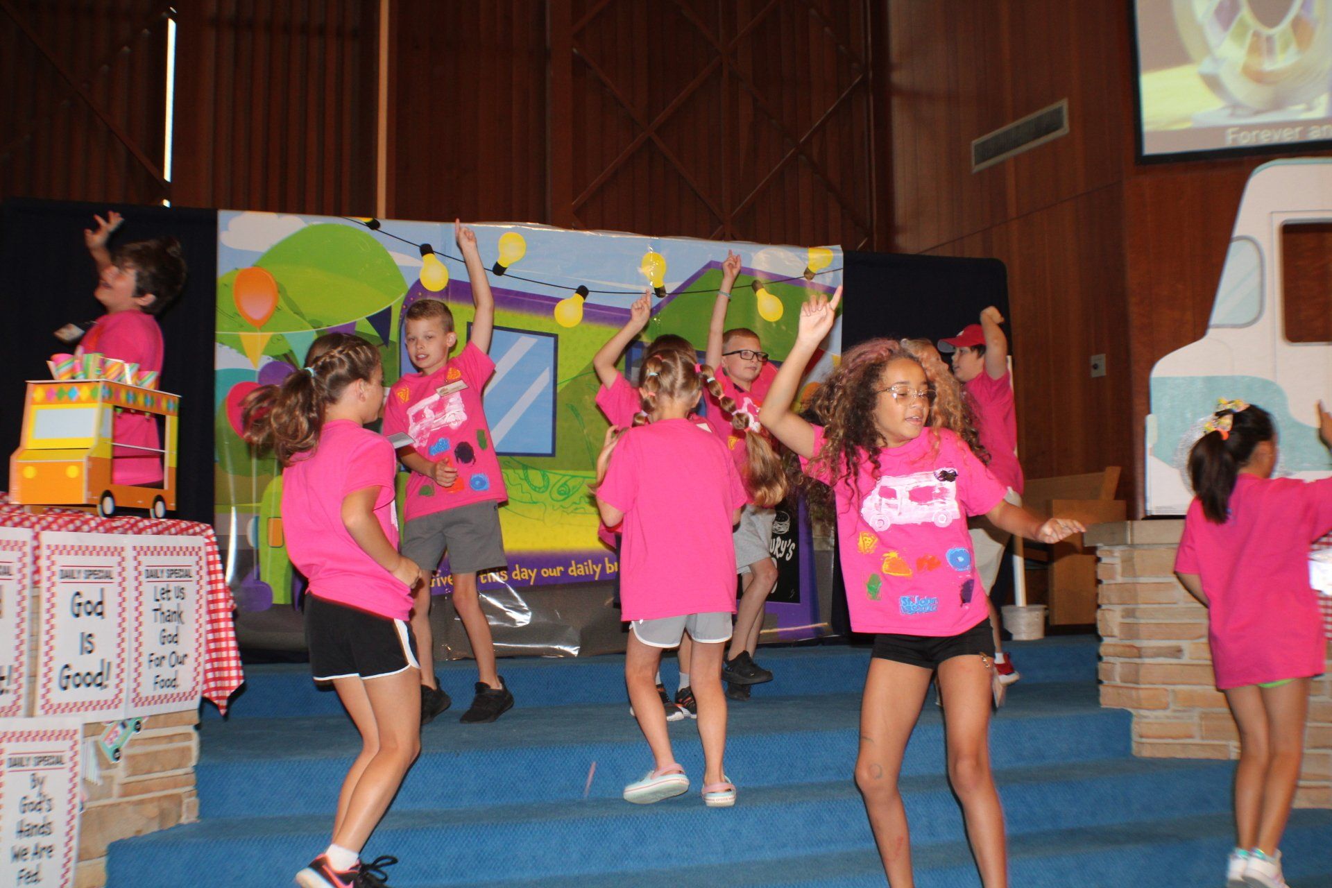 Children in pink shirts performing a dance on a stage with colorful props and a festive backdrop