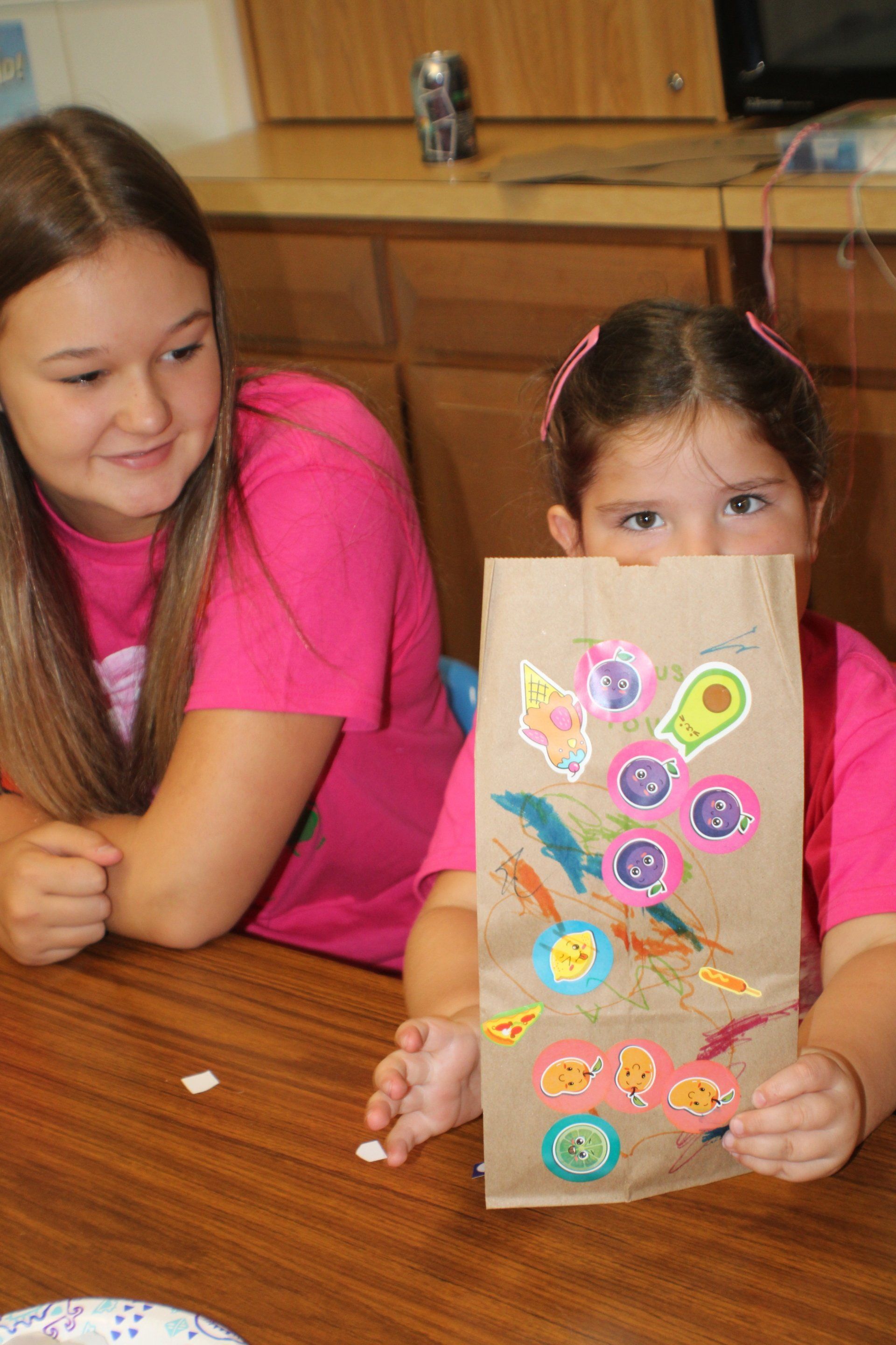 Two children at a table, one holding a decorated paper bag with colorful stickers.