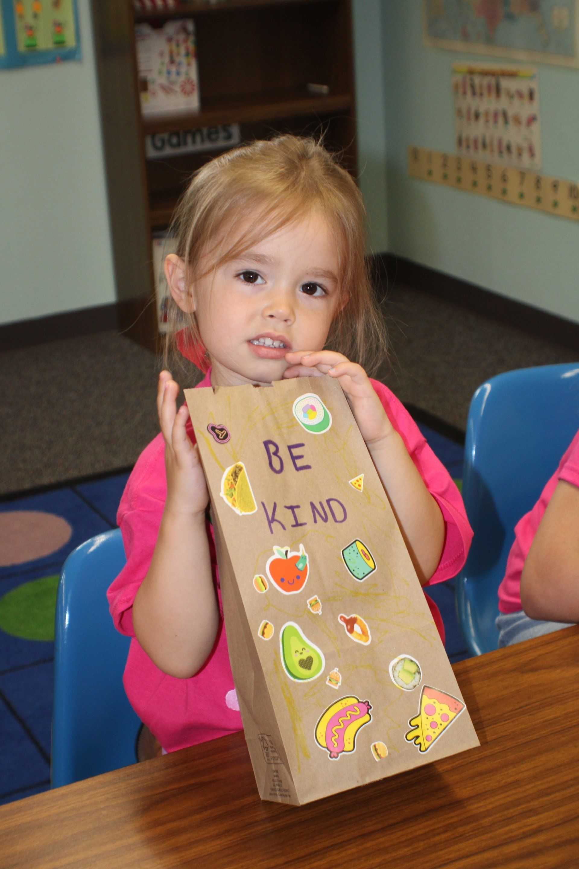 Child holding a decorated paper lunch bag at a classroom table, with “BE KIND” written on it.