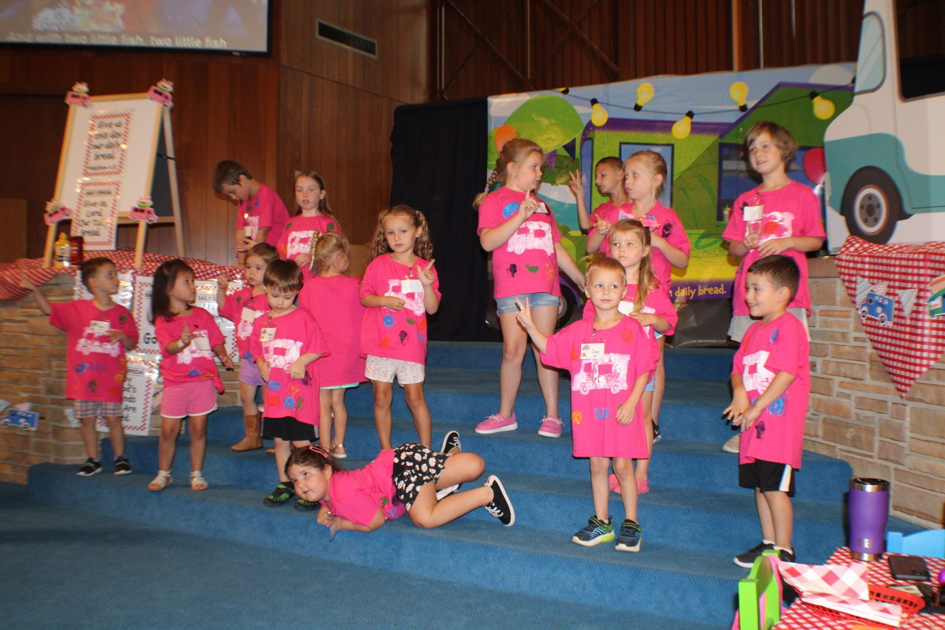 Children in pink shirts perform on a stage with a colorful bus backdrop and blue carpet
