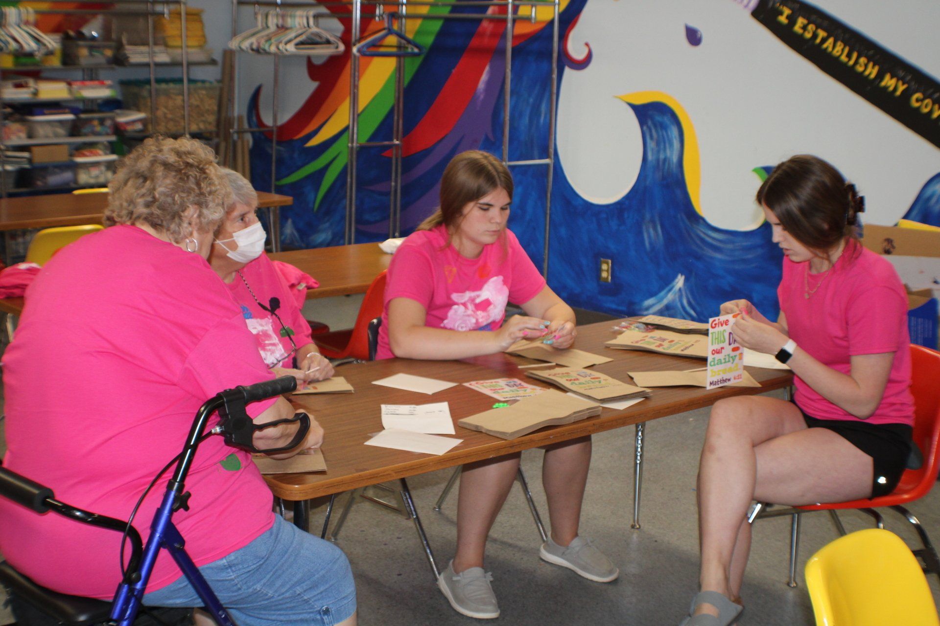 Three people in pink shirts sit at a table crafting with paper and tools in a colorful room.