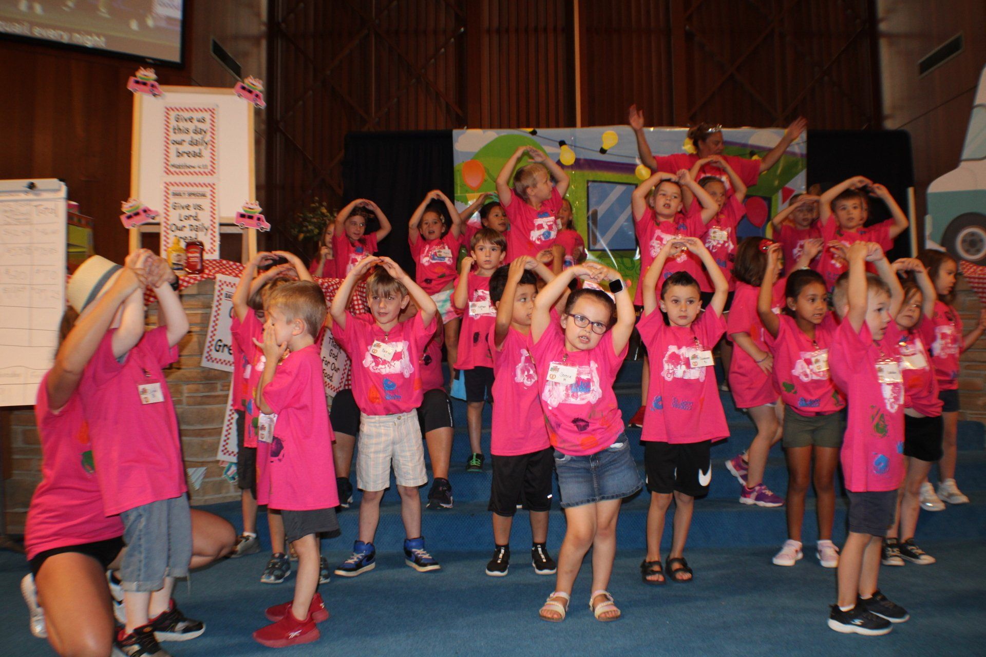Children in pink shirts posing on a stage, many making hand-heart shapes