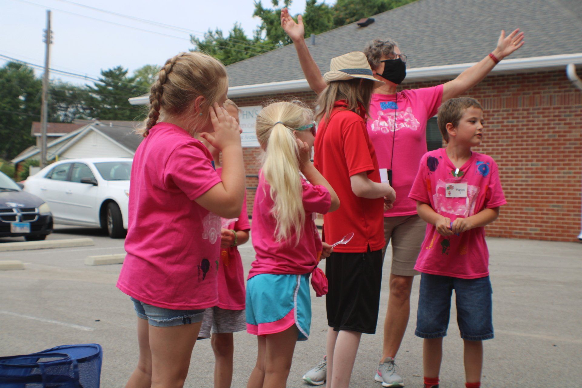 Children in pink shirts high-five outdoors near a brick building and parked cars