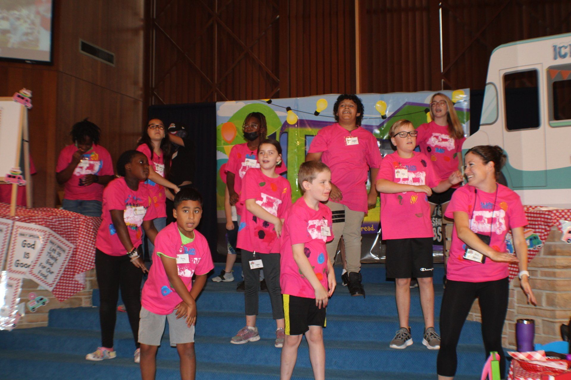 Children in pink shirts performing on a stage with microphones and a colorful backdrop