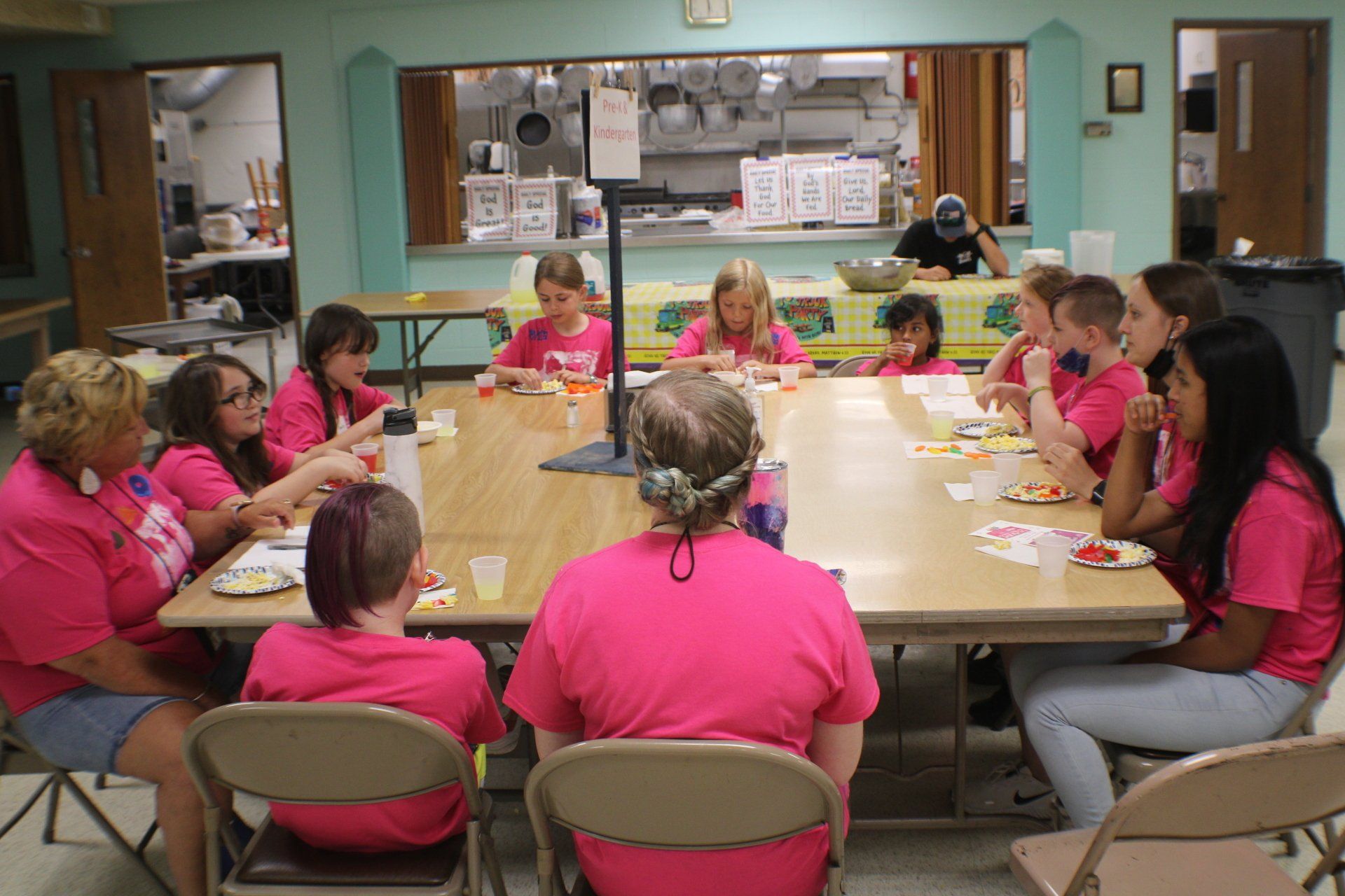 Children in pink shirts sit around a cafeteria table, eating snacks and talking.