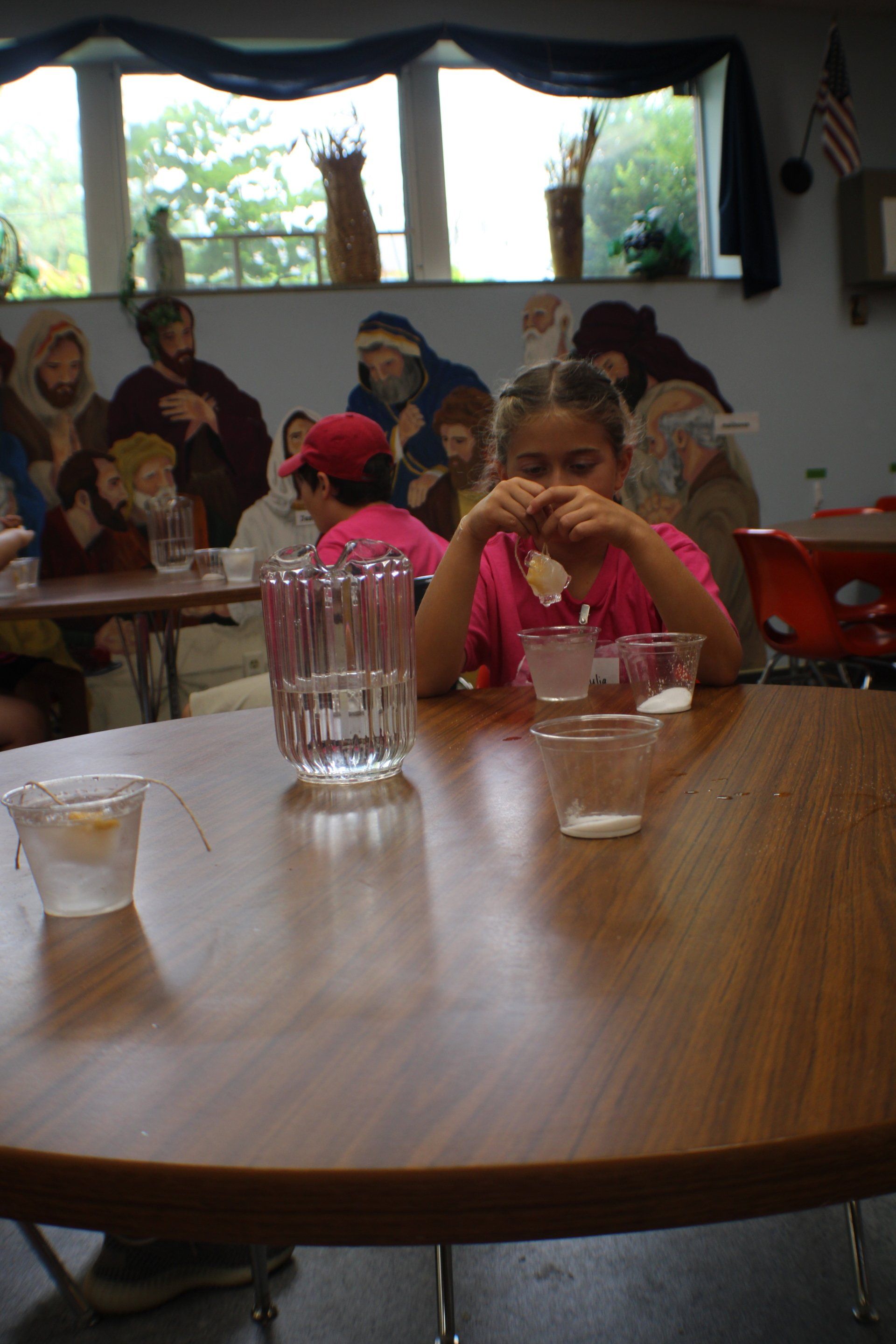 Child in pink shirt sitting at a table in a classroom, with cups and a glass pitcher in the foreground.