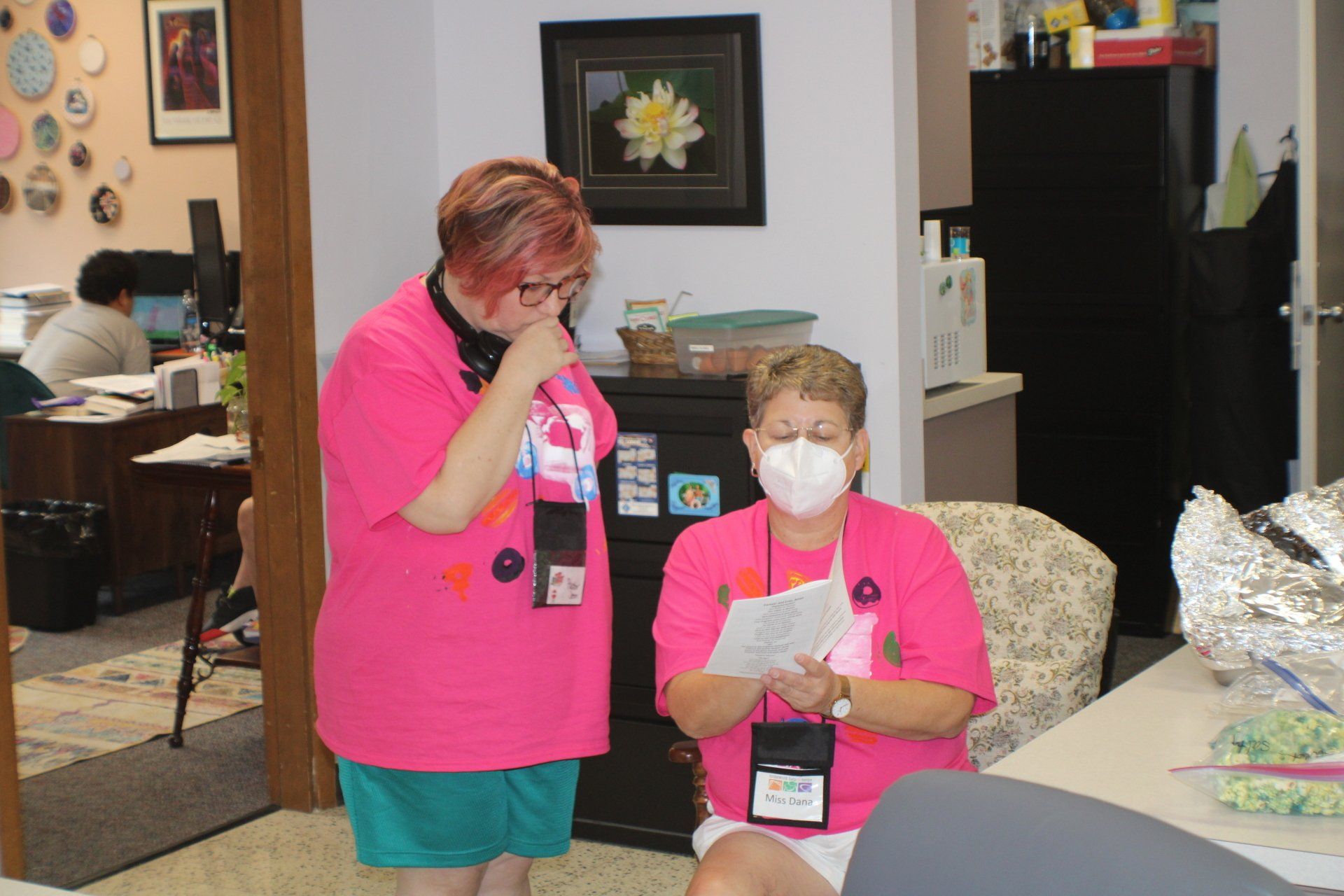 Two masked people in pink shirts at a table in a room, one standing and one seated holding a paper.