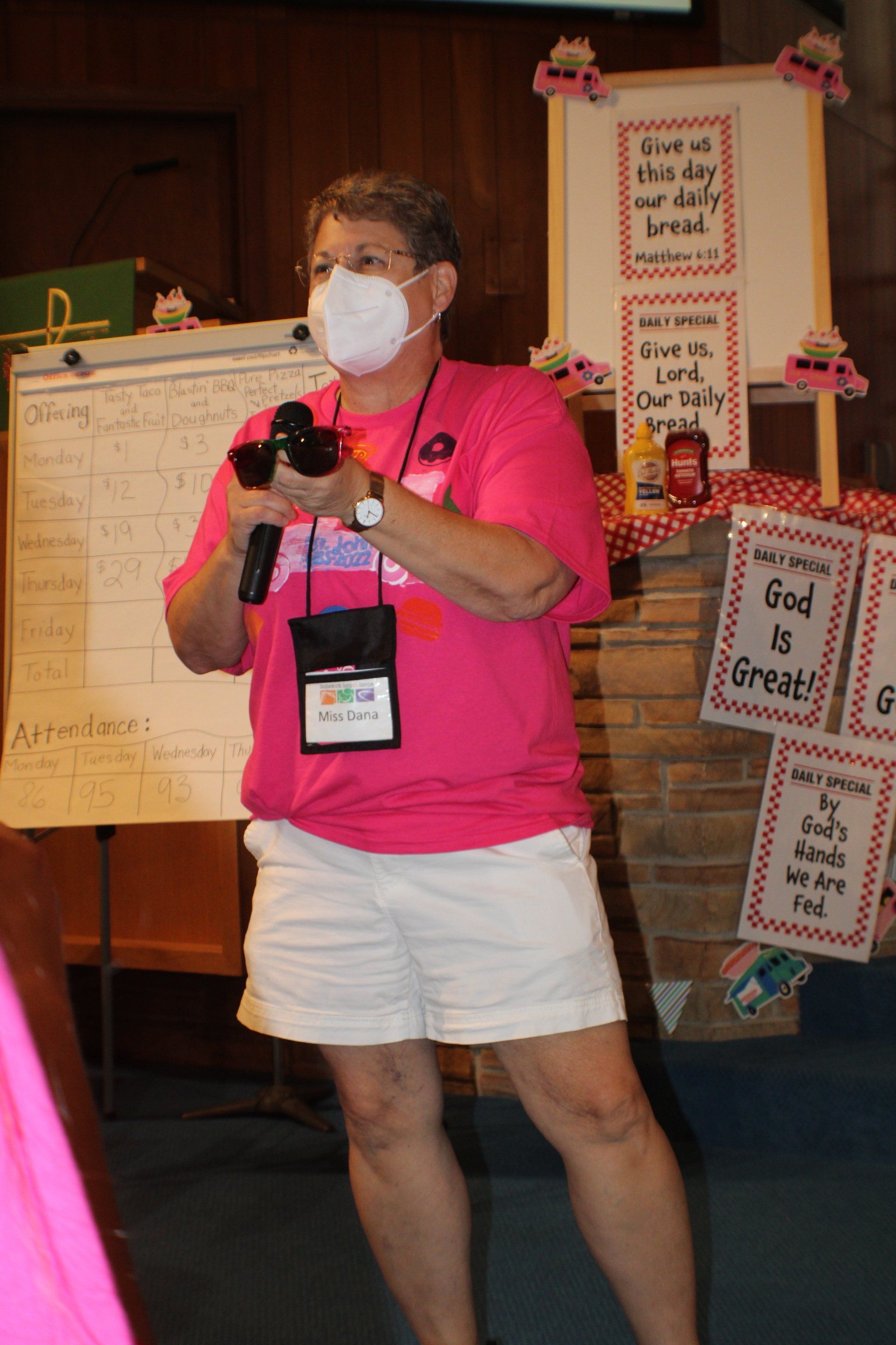 Masked speaker in pink shirt and white skirt holding a microphone at an indoor event with posters.