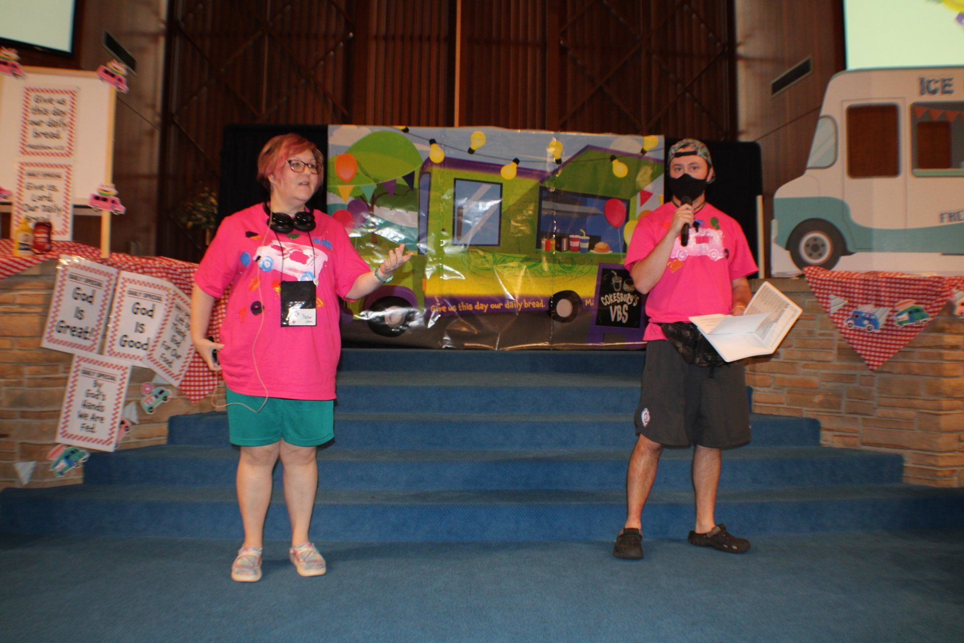 Two people in pink shirts standing on a stage in front of a colorful display and props