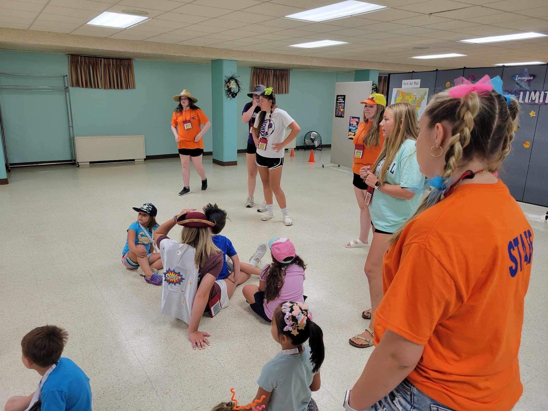 Children and adults in colorful shirts crouch in a dance studio, watching a group activity.