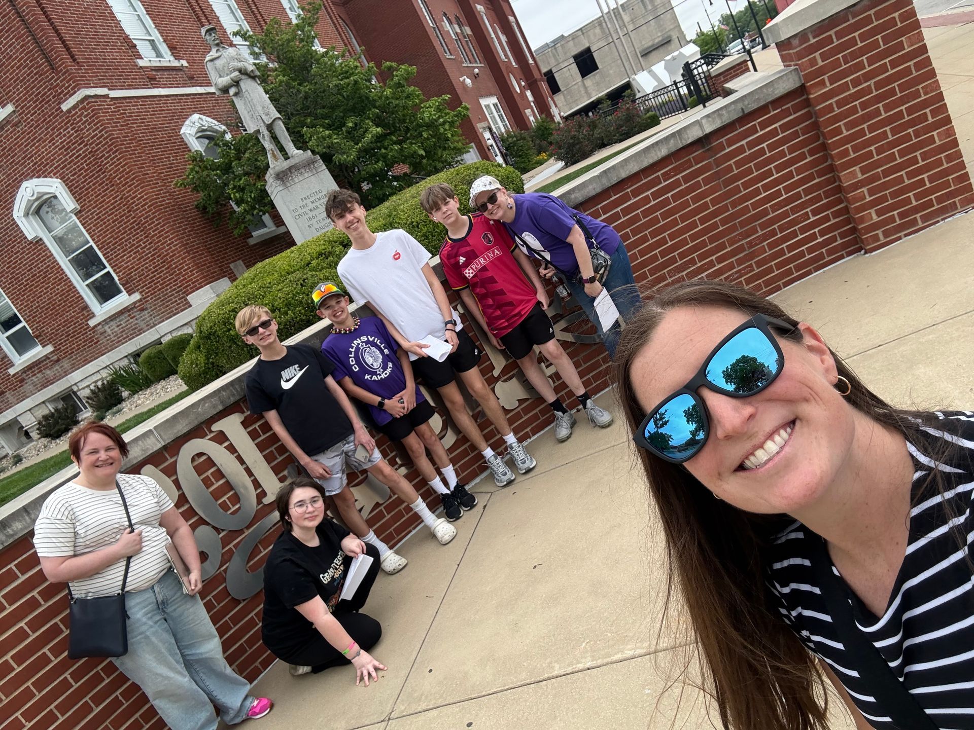 Group posing outdoors on a campus sidewalk, smiling in front of a brick building and fountain.