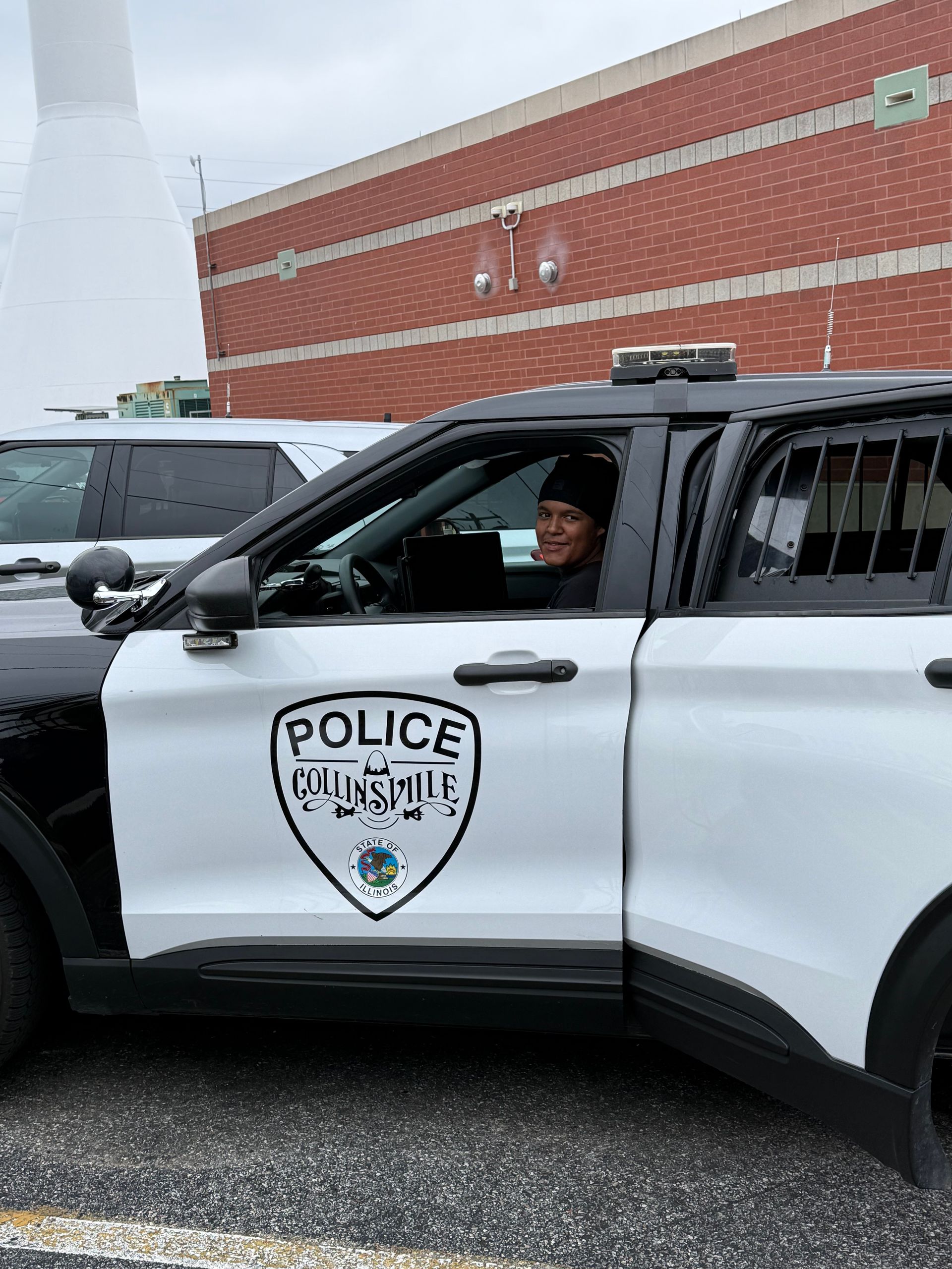 Police officer in a black-and-white patrol SUV beside a brick building, officer smiling from the open window