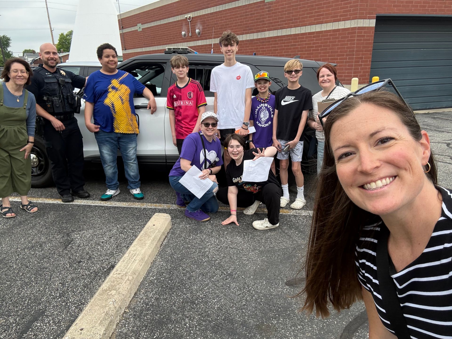 Group posing outside a brick building; smiling woman takes a selfie in the foreground.