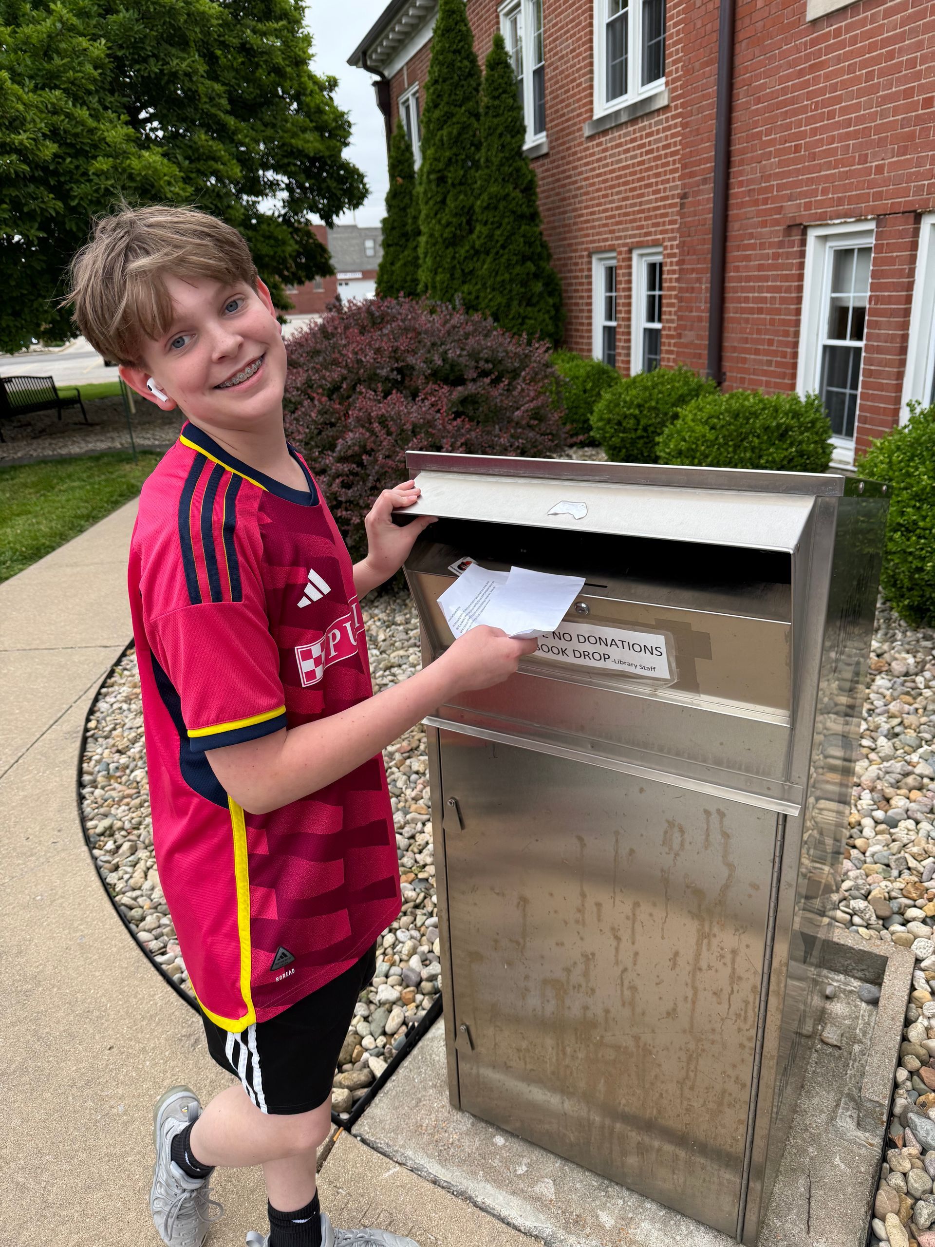 Smiling person in a red sports jersey posts a letter into a metal mailbox outside a brick house.