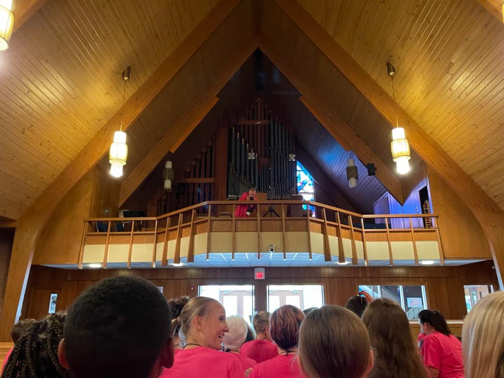 People seated in a wooden church hall facing a balcony and organ pipes at the front