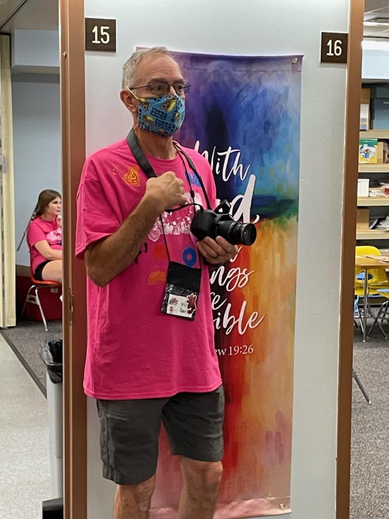 Man in pink shirt and mask posing in a library between aisles, holding a camera with colorful banner behind him