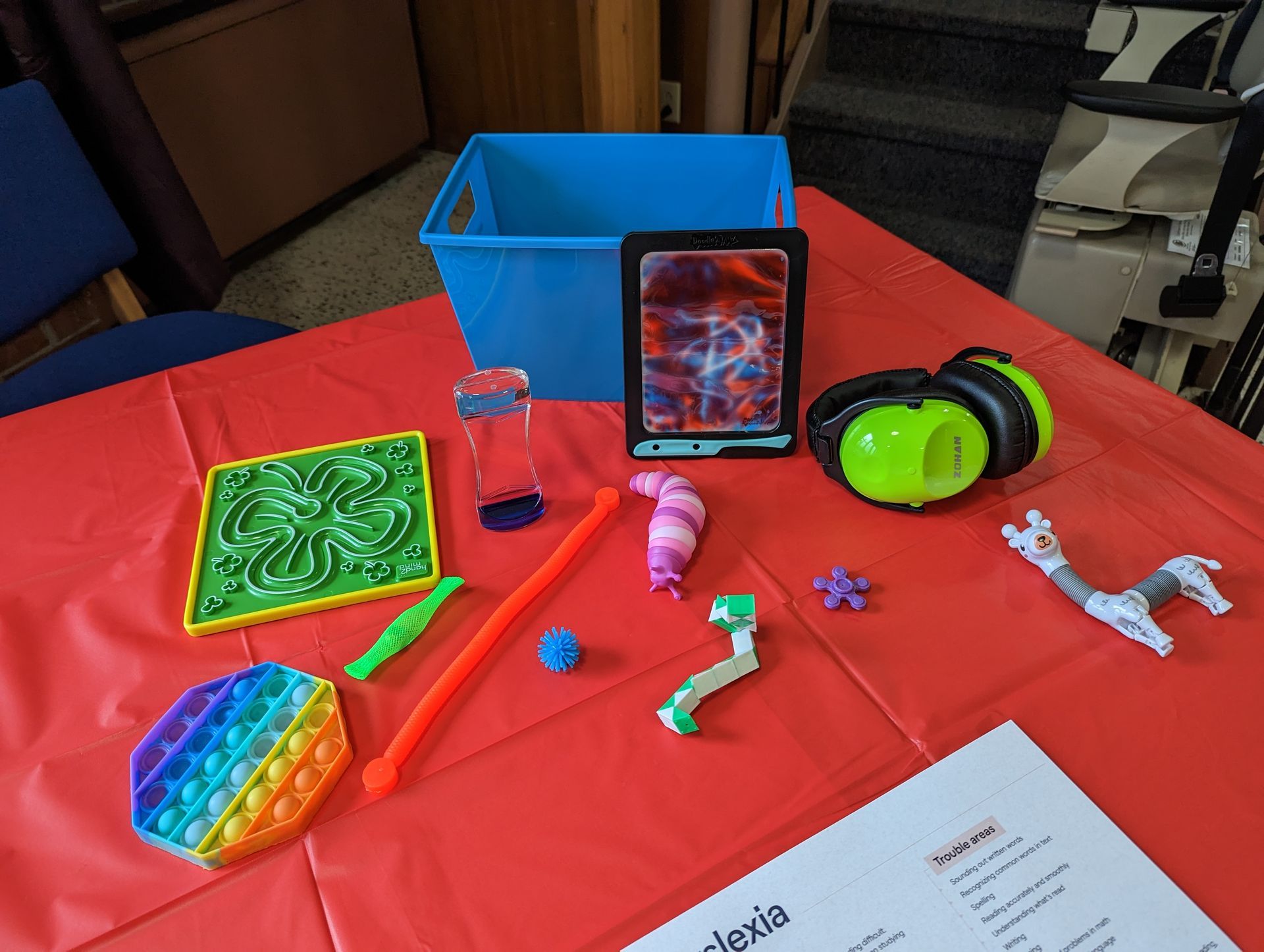 Bright red table with assorted colorful fidget toys, headphones, and a blue storage bin at the back