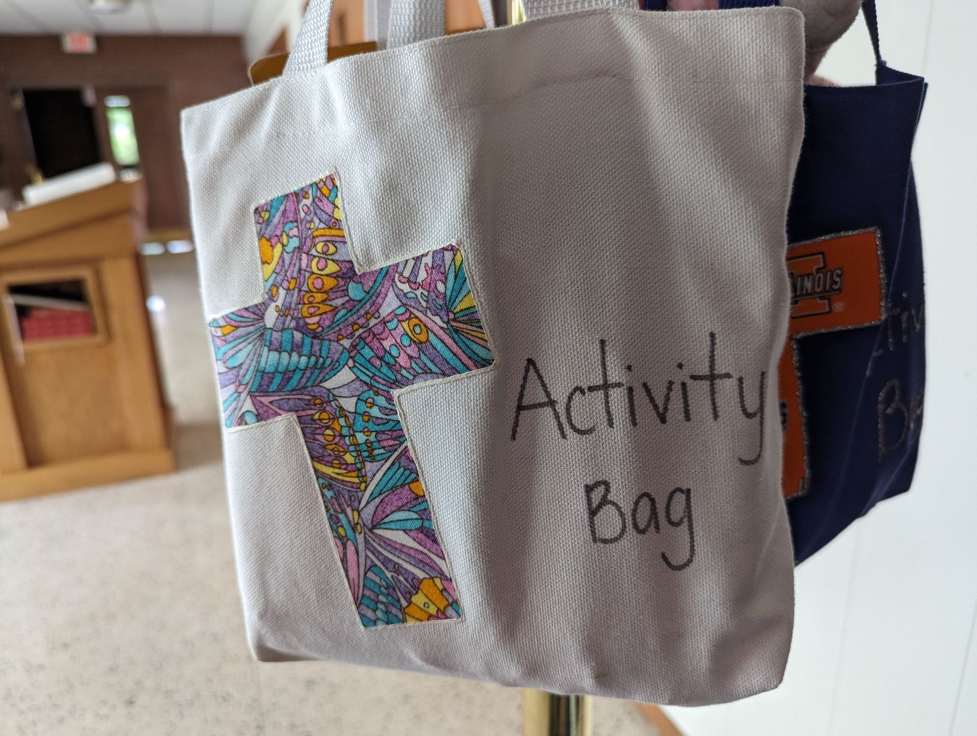 White activity bag with colorful cross and “Activity Bag” text hanging indoors