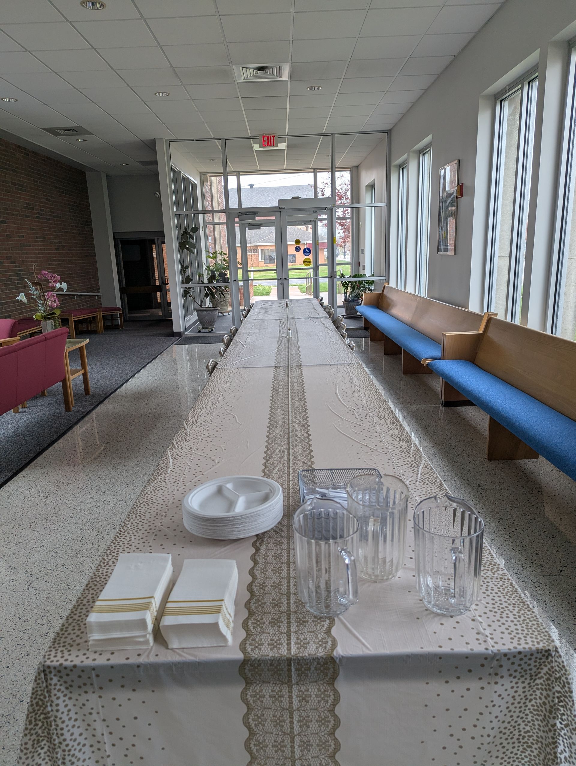 Indoor hallway with a long table set with plates, cups, and napkins, flanked by benches and windows.