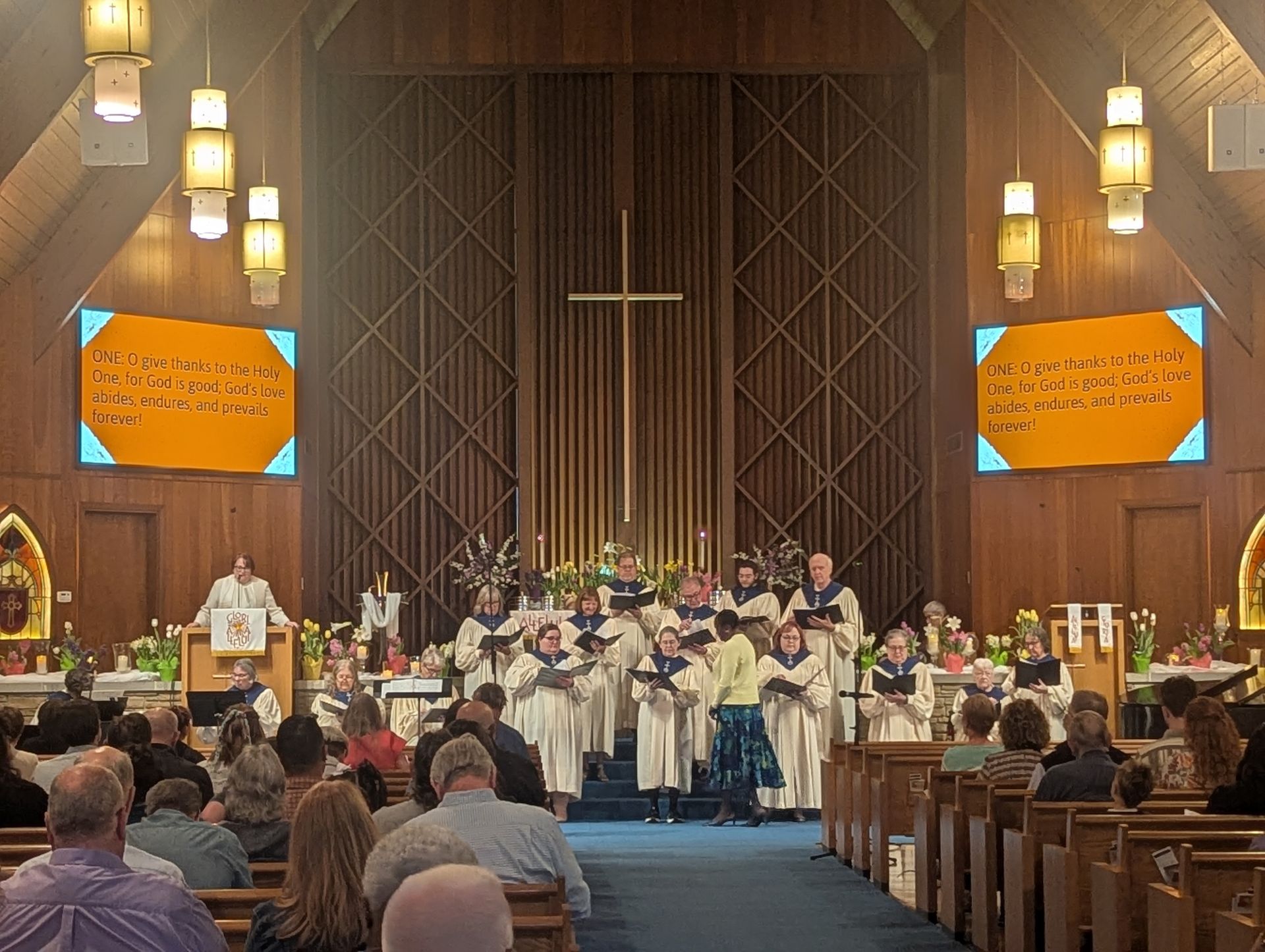 Church choir performing in a wood-paneled sanctuary, with congregation seated and orange hymn boards on walls