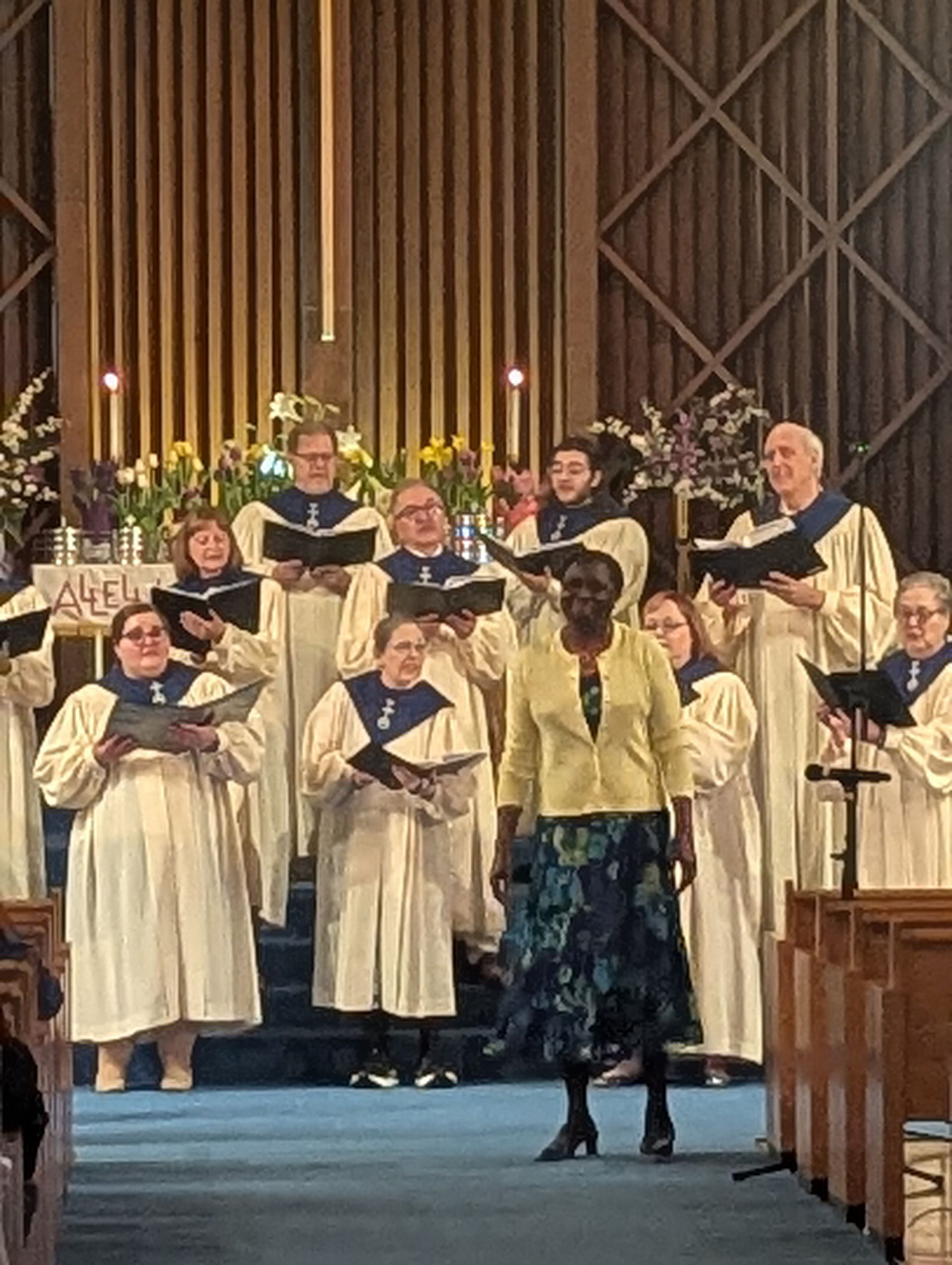 Choir in white robes singing on a church stage, with a soloist in front and wooden pews at right.