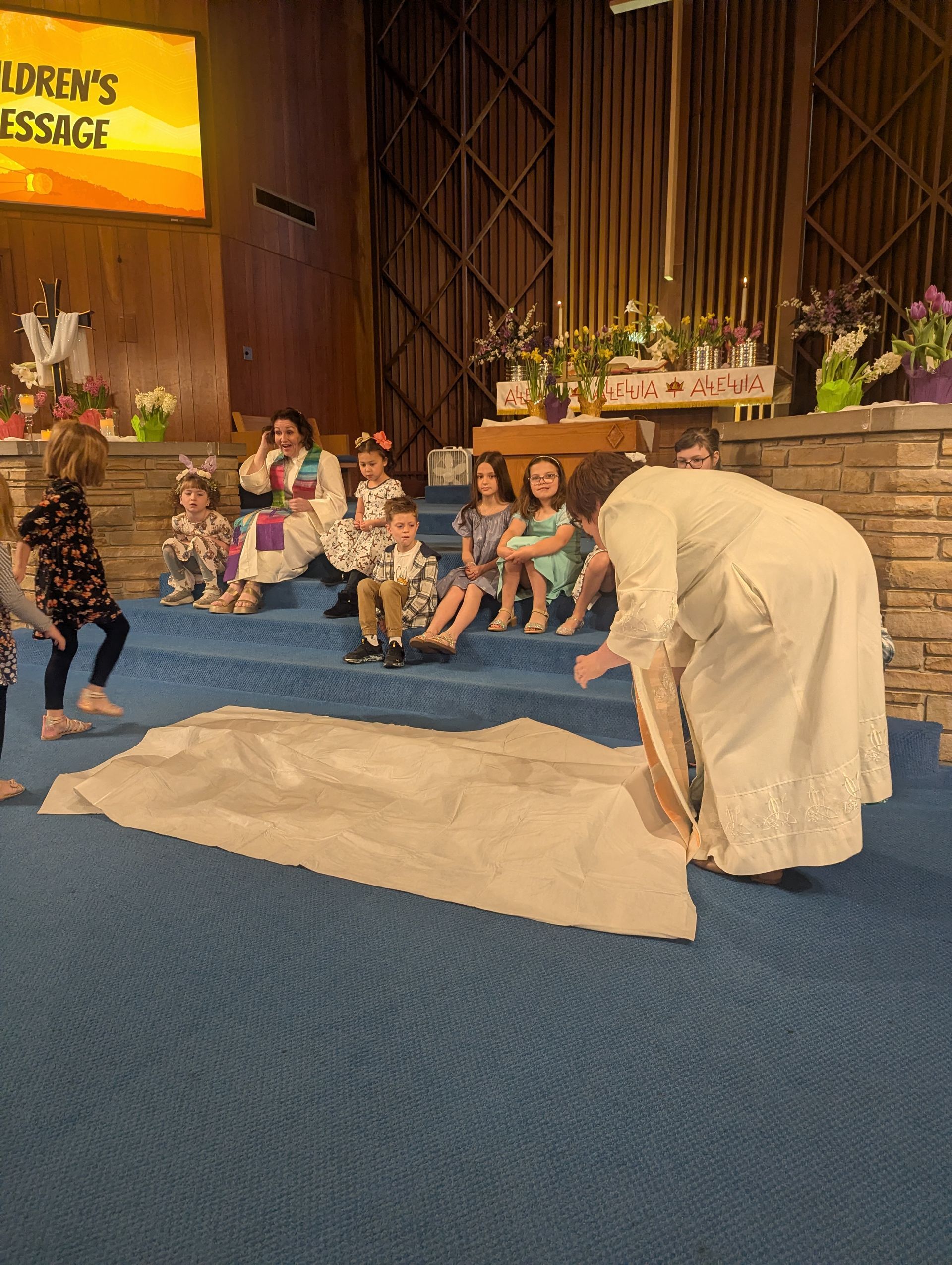People seated on a blue-carpeted stage near a white-draped table during a church service