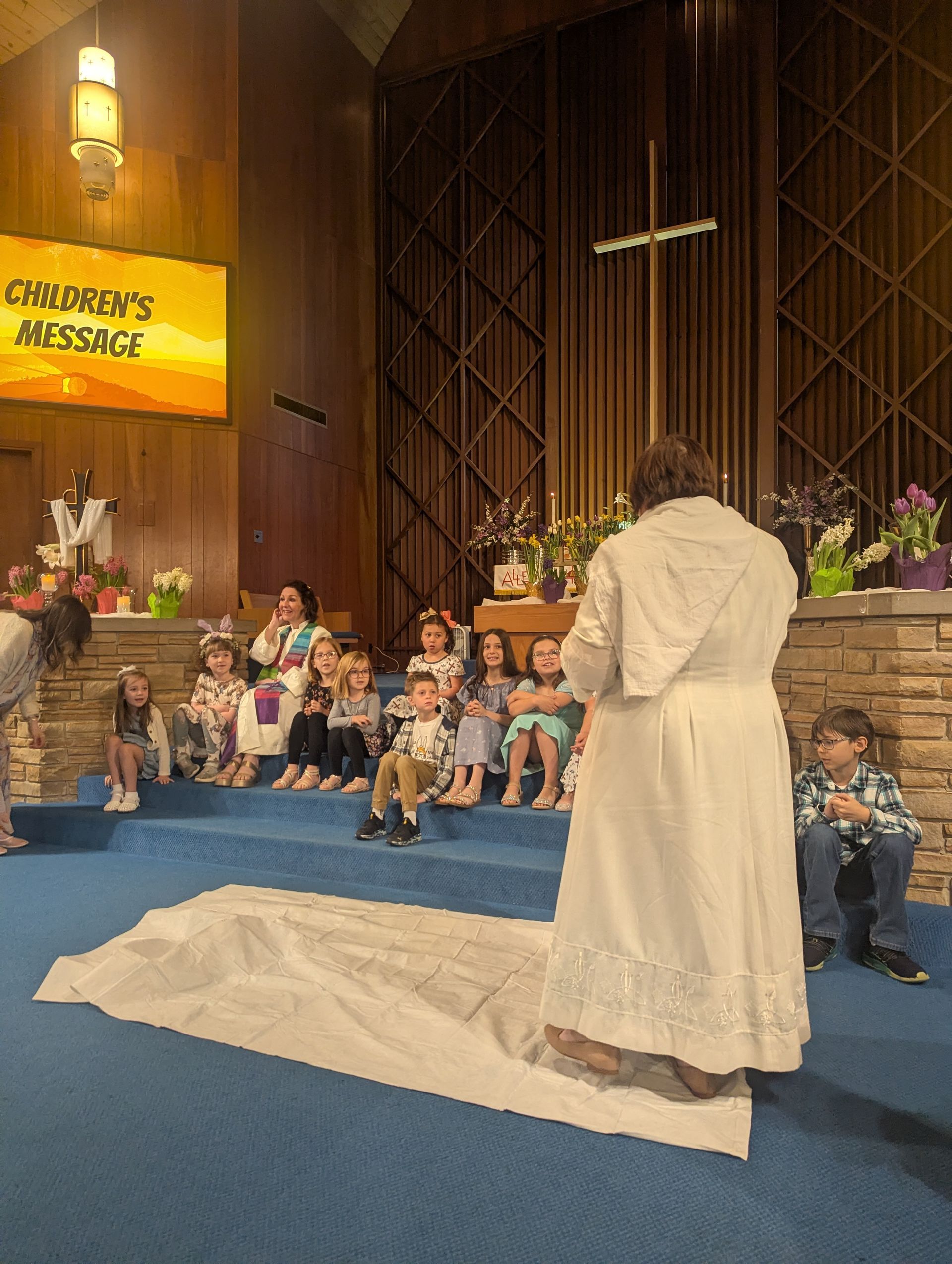 Person in white robe stands before seated children in a church sanctuary with blue carpet and candles.