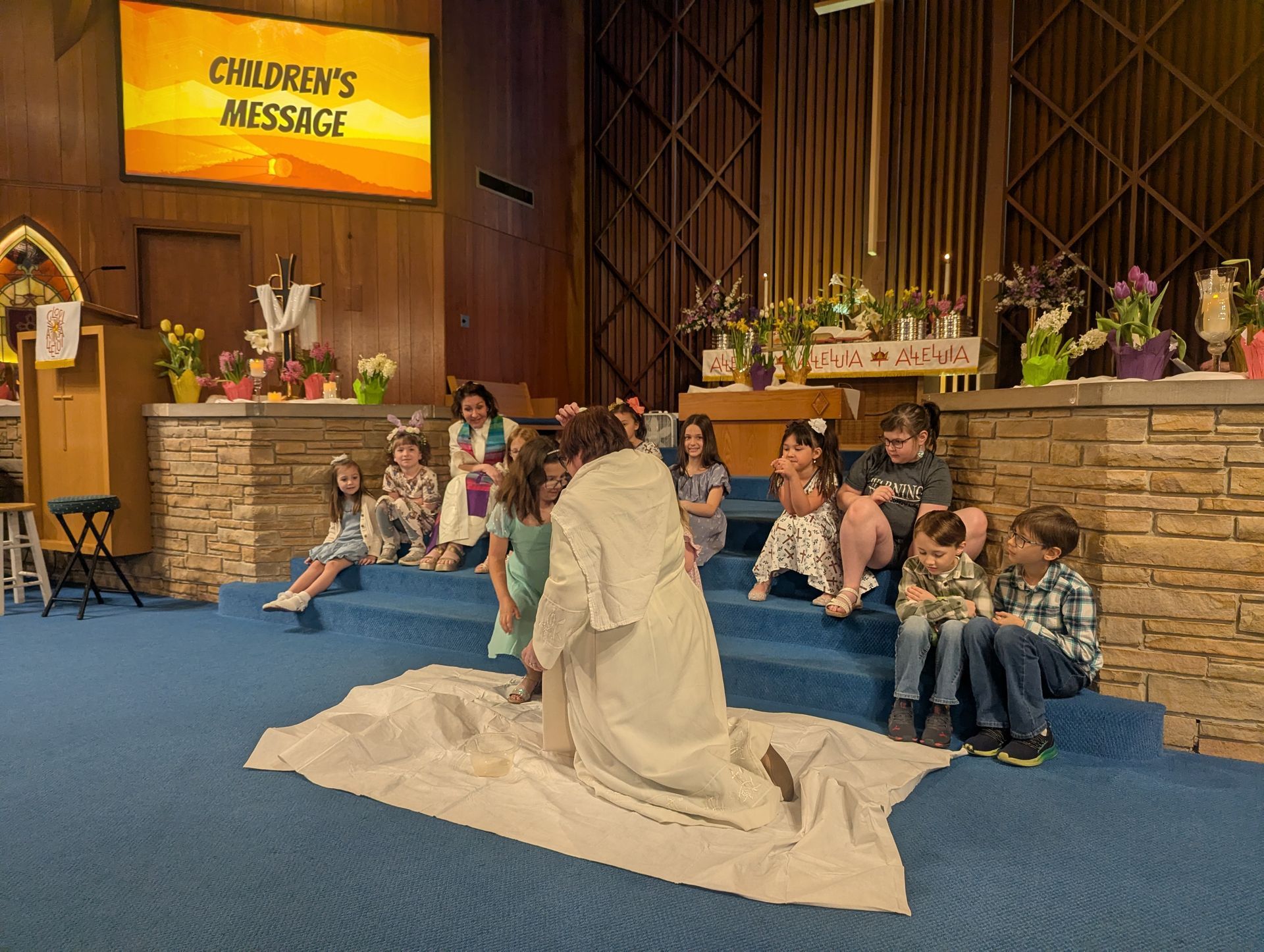 Group seated on blue carpet in a church, facing a draped figure near a sign reading “Children’s Message.”