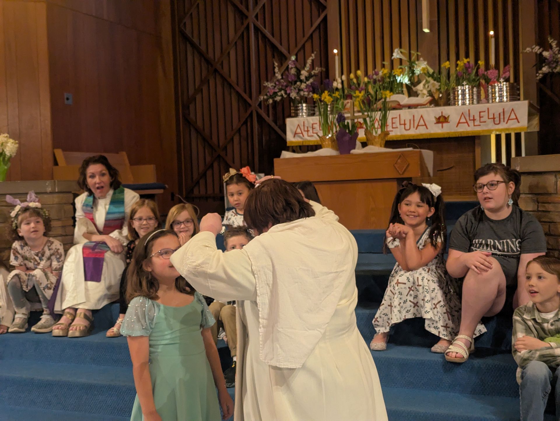 Children seated on a church stage while an adult kneels in front of them during a ceremony