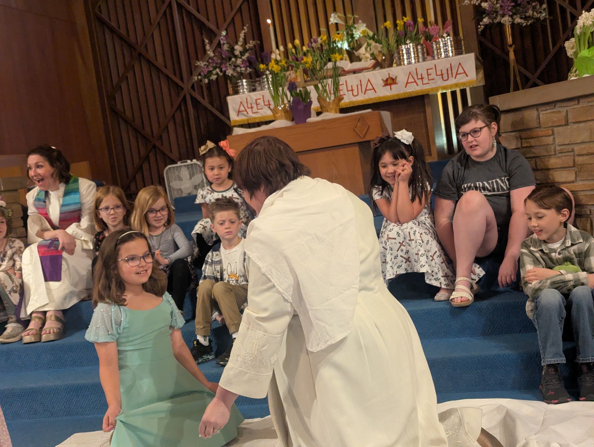 Woman in white kneels before children seated on blue steps in a decorated indoor stage setting