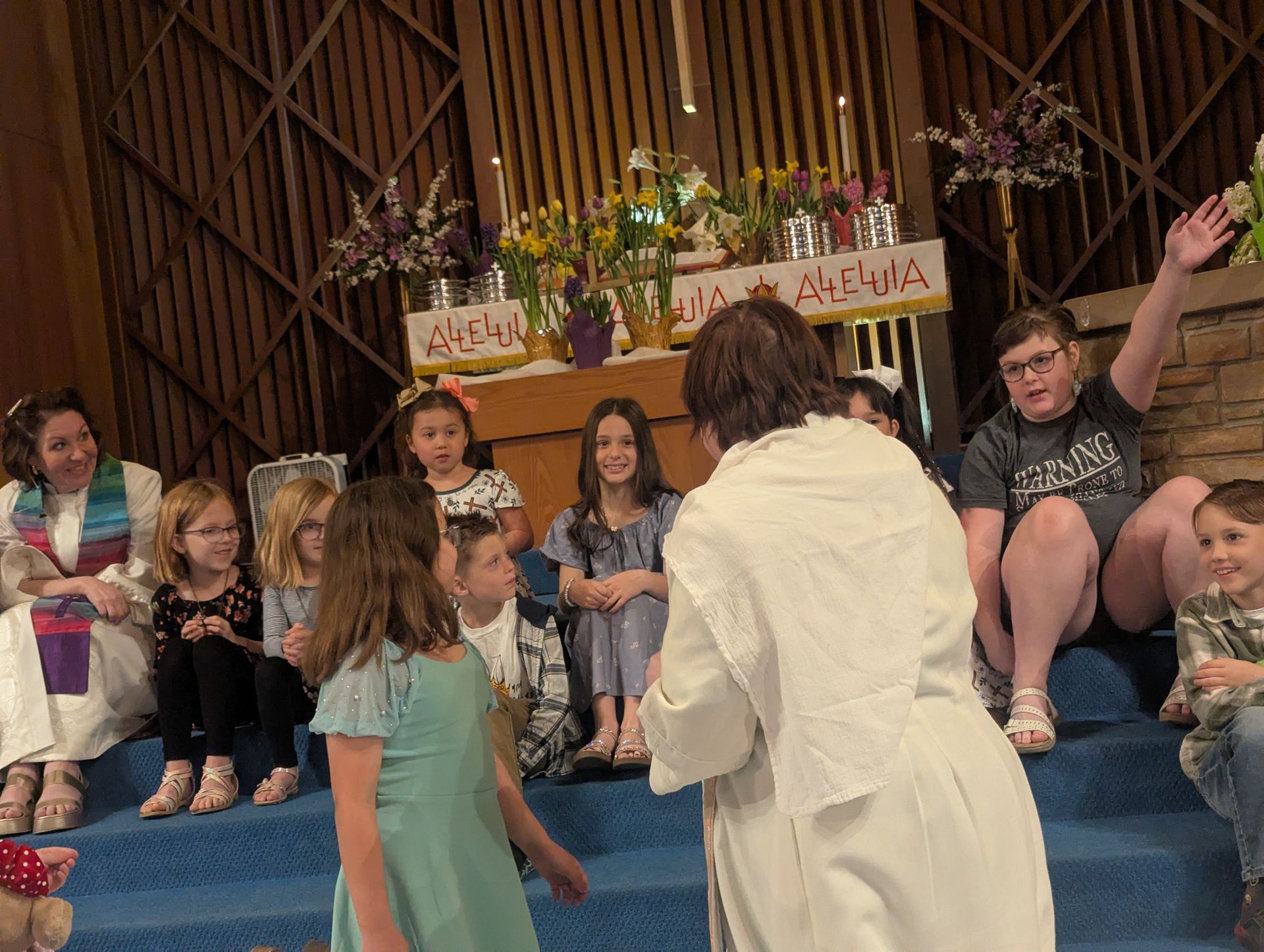 Children seated on blue carpet watch a woman in white in a church-like setting