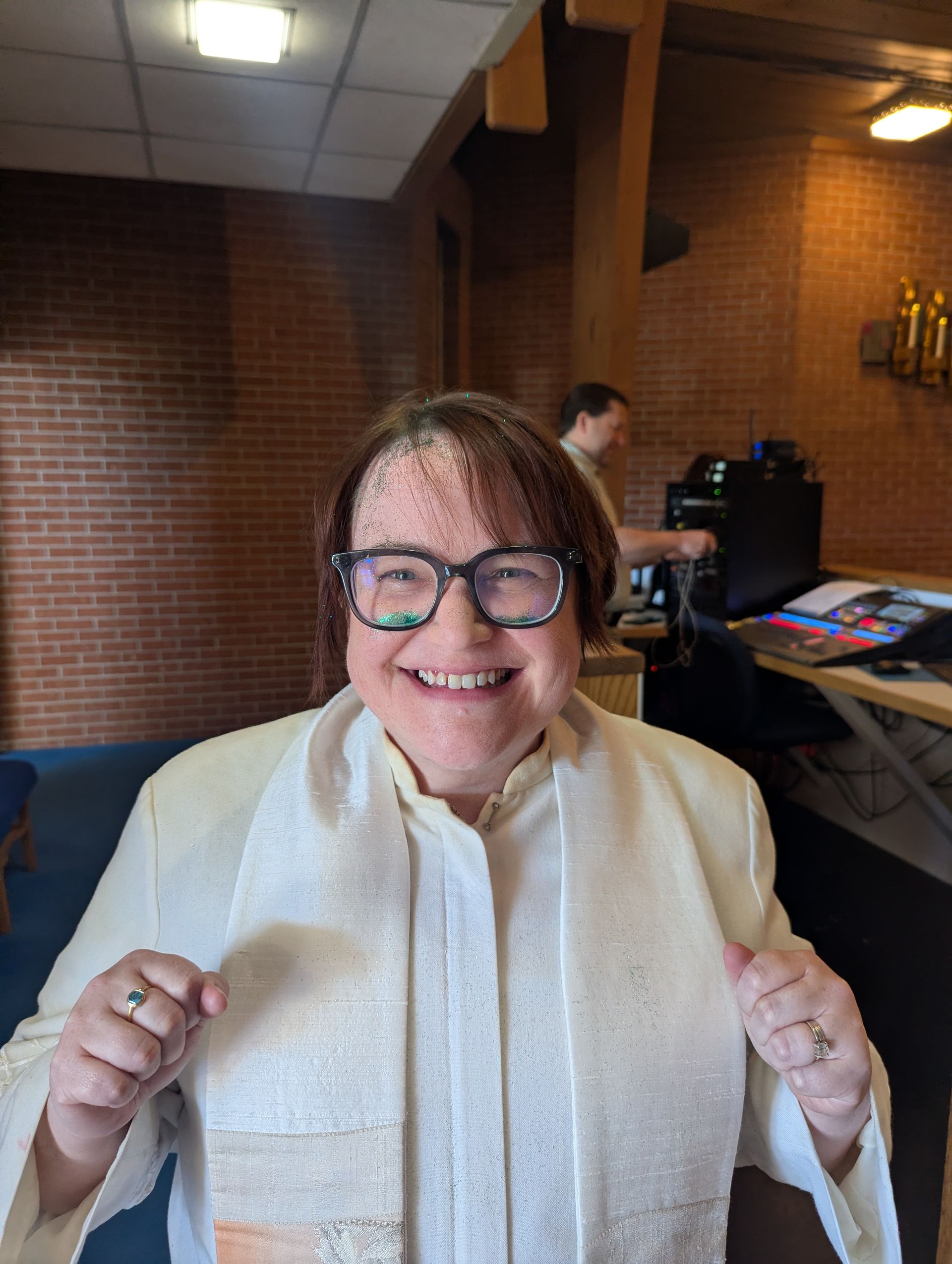 Smiling person in glasses and white sweater vest, standing indoors against a brick wall.