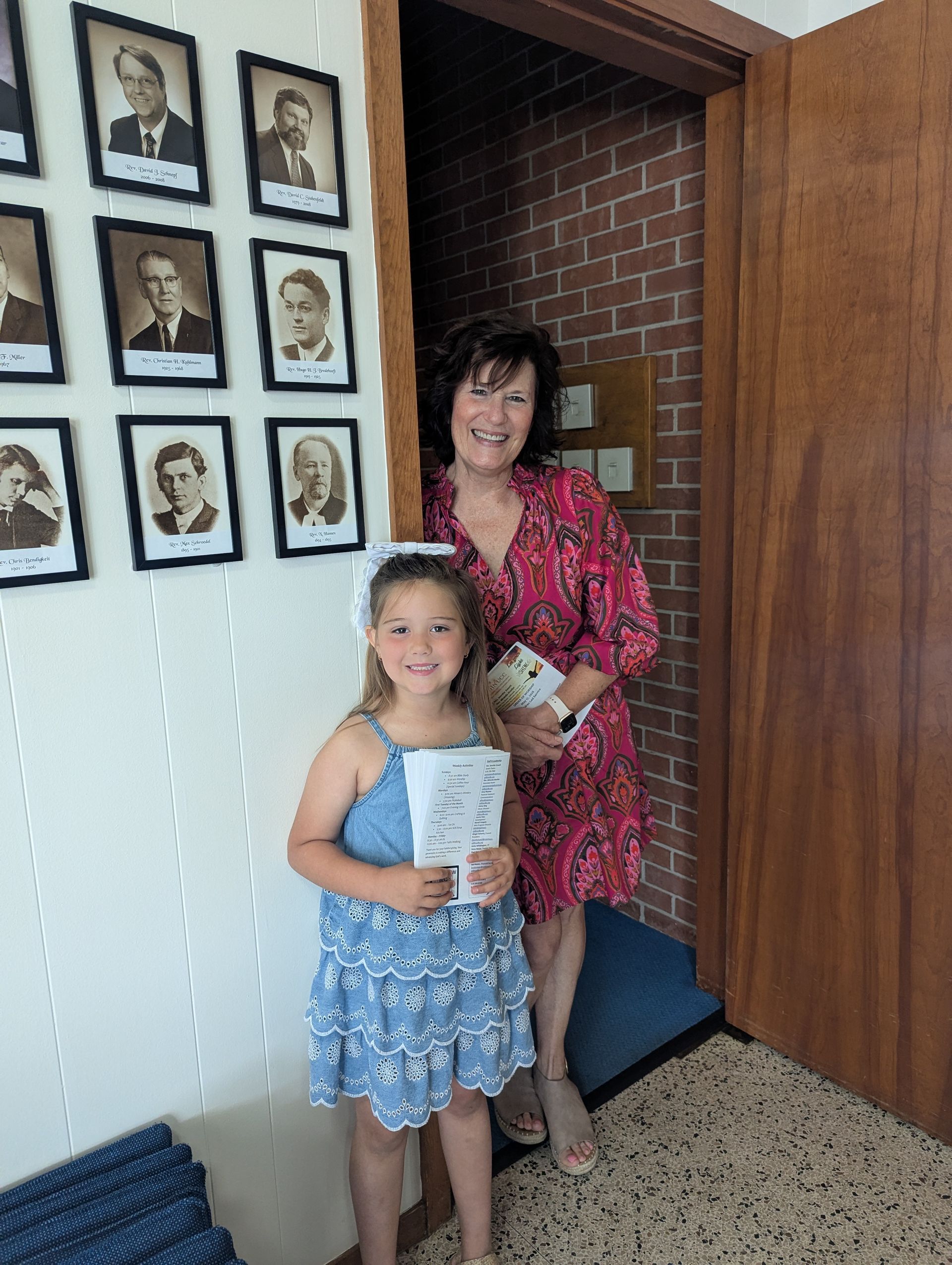 Woman and child standing indoors by framed photos, holding papers and a small stuffed toy.