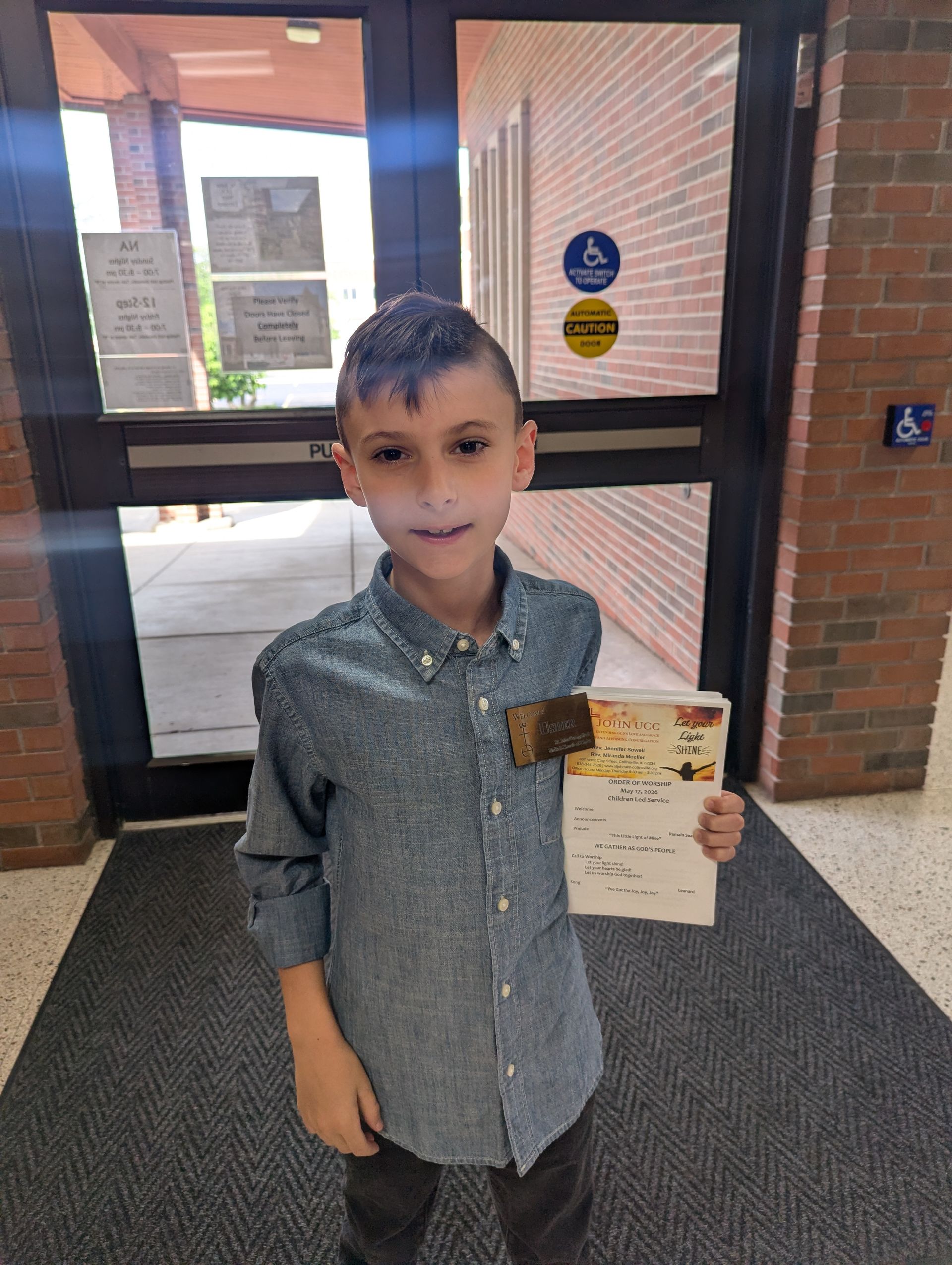 Child standing indoors by a glass door, holding a certificate or award.