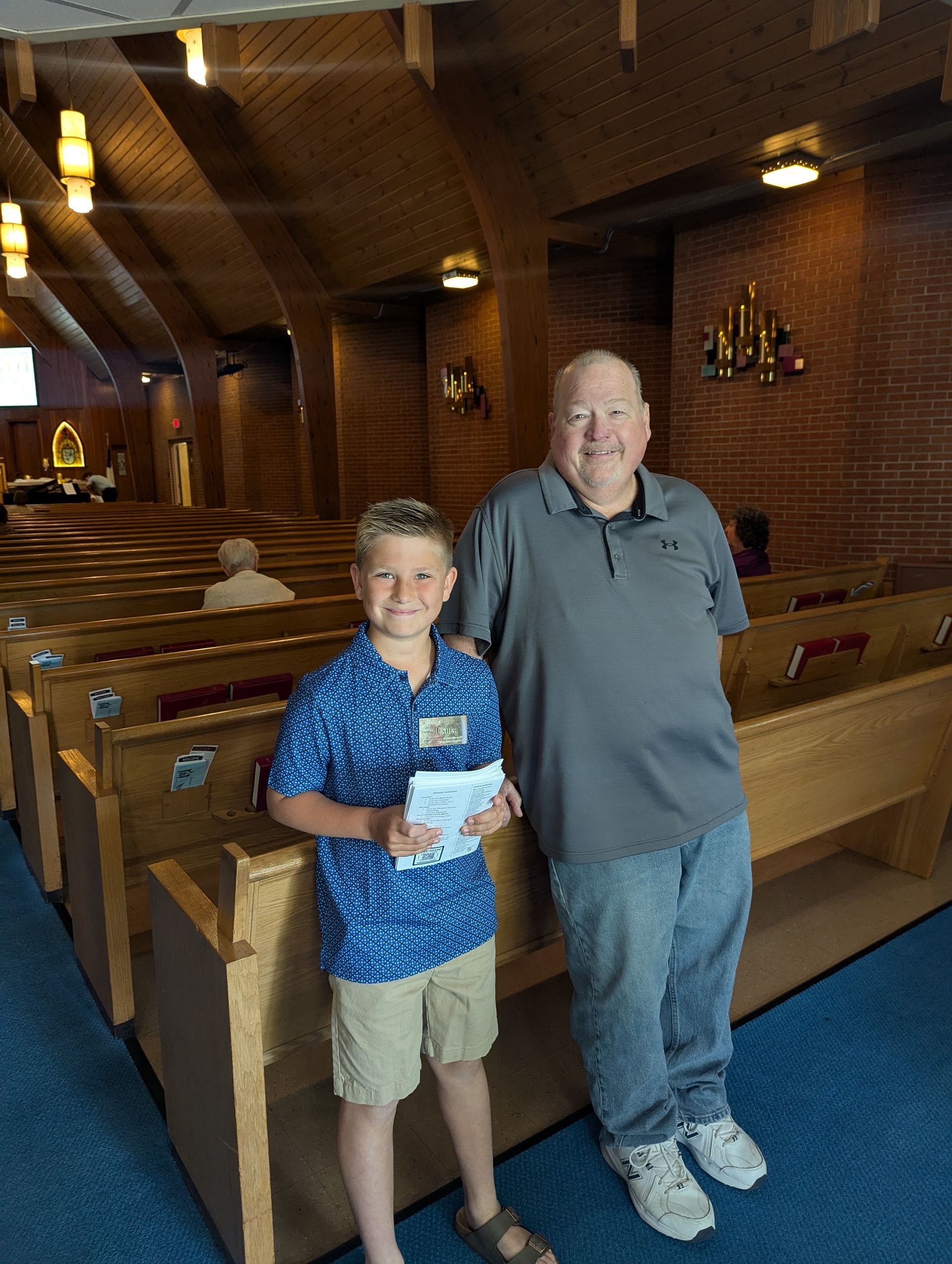 Two people standing in a wooden church aisle, one holding a paper, under warm ceiling lights.