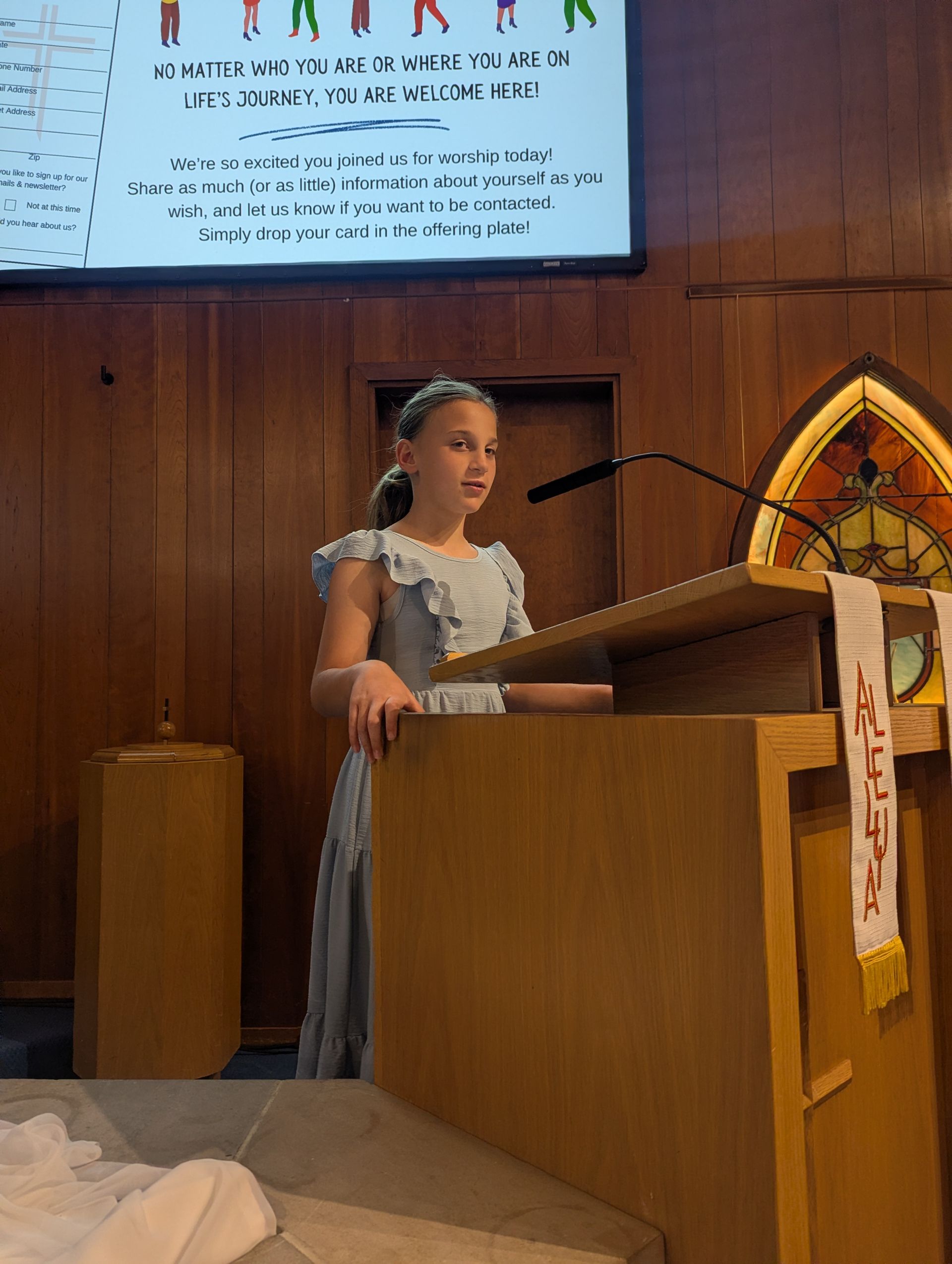 Girl standing at a church lectern, speaking into a microphone under a projection screen.