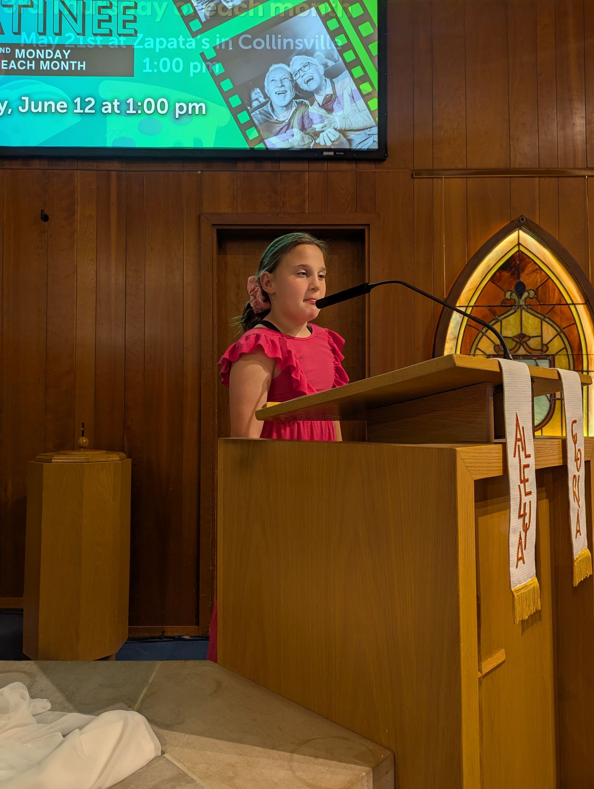 Person in a pink dress speaking at a wooden lectern in a church-like hall, with a screen behind them.
