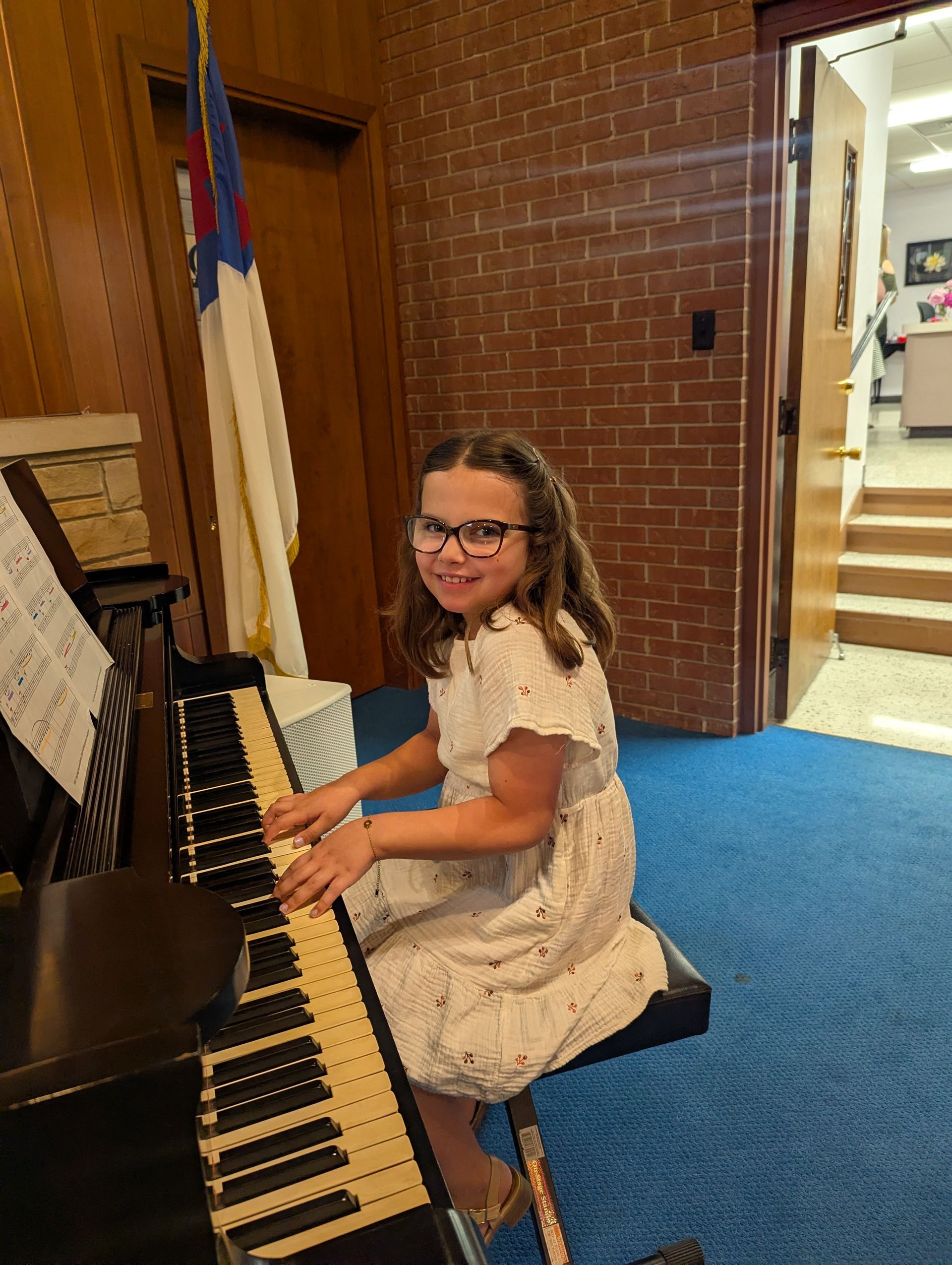 Girl playing a black piano in a church, smiling beside a flag and brick wall.