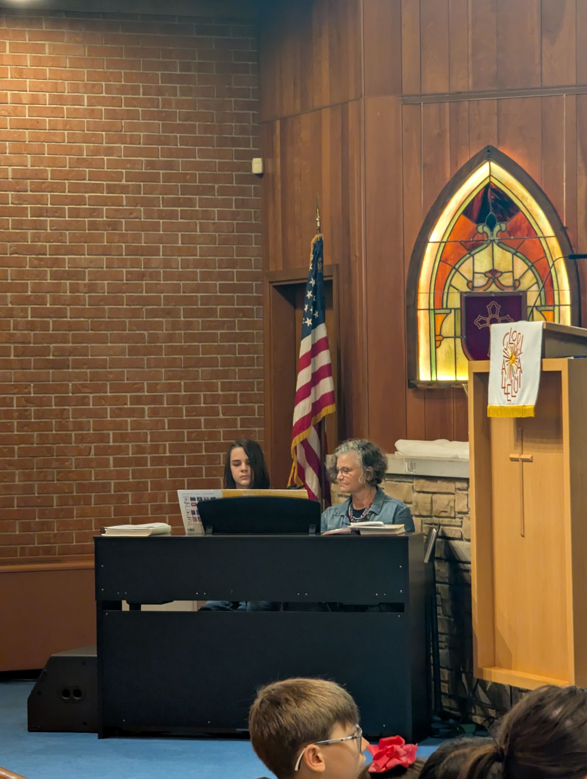 Two people seated at a piano in a church, with an American flag, lectern, and stained-glass window nearby.
