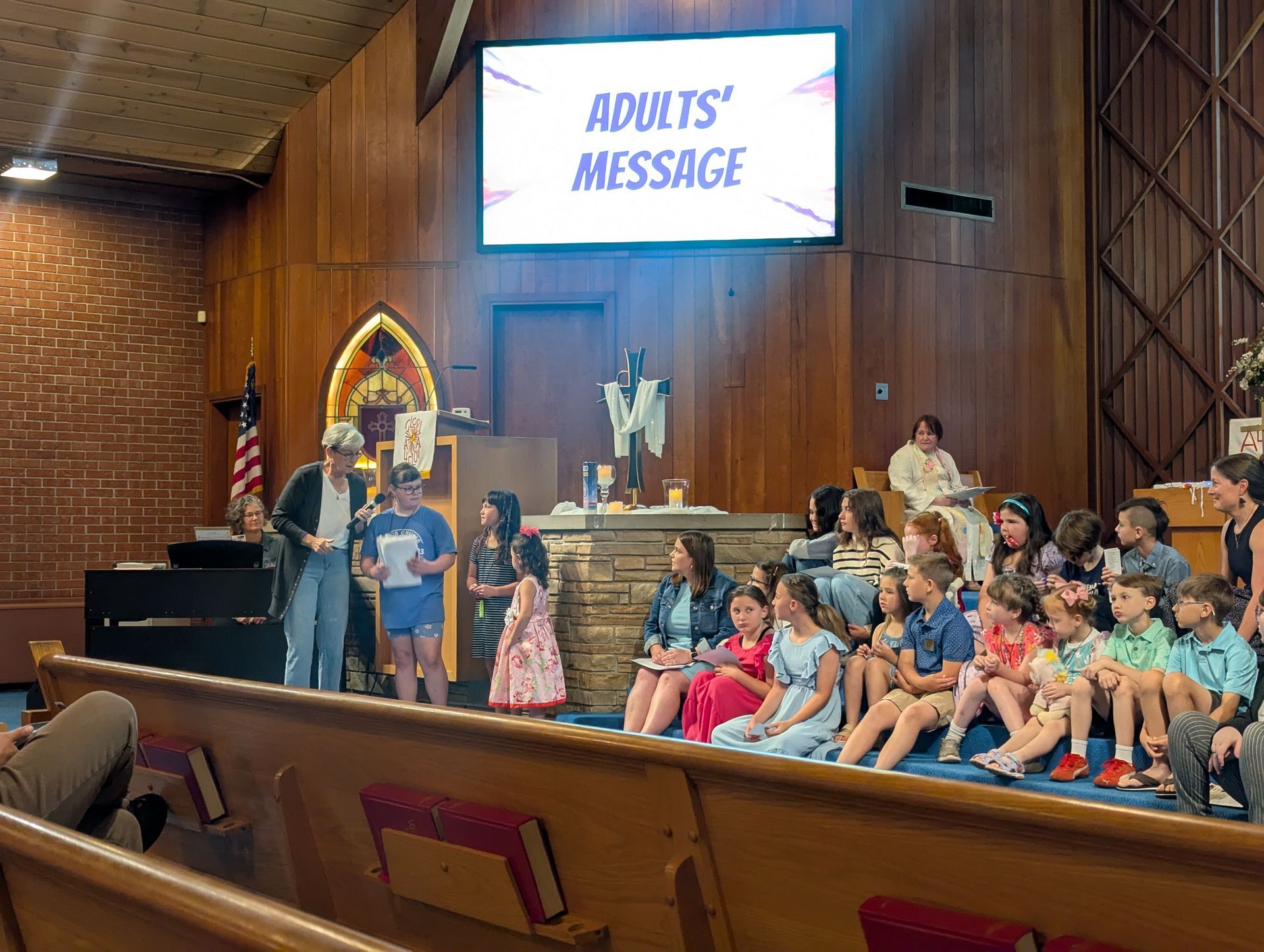 Children seated in a church sanctuary watching a stage sign reading “ADULTS’ MESSAGE”
