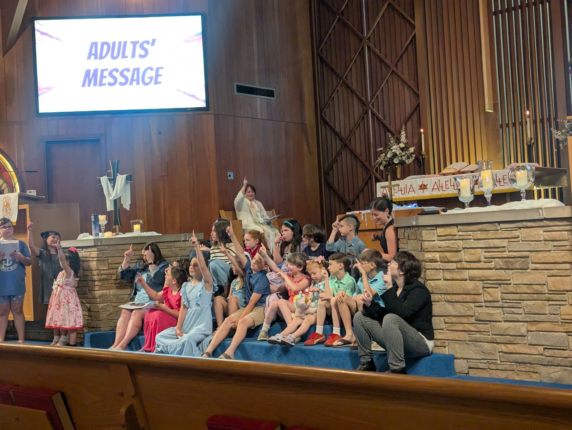 Children seated in a church sanctuary beneath a screen reading “ADULTS’ MESSAGE”